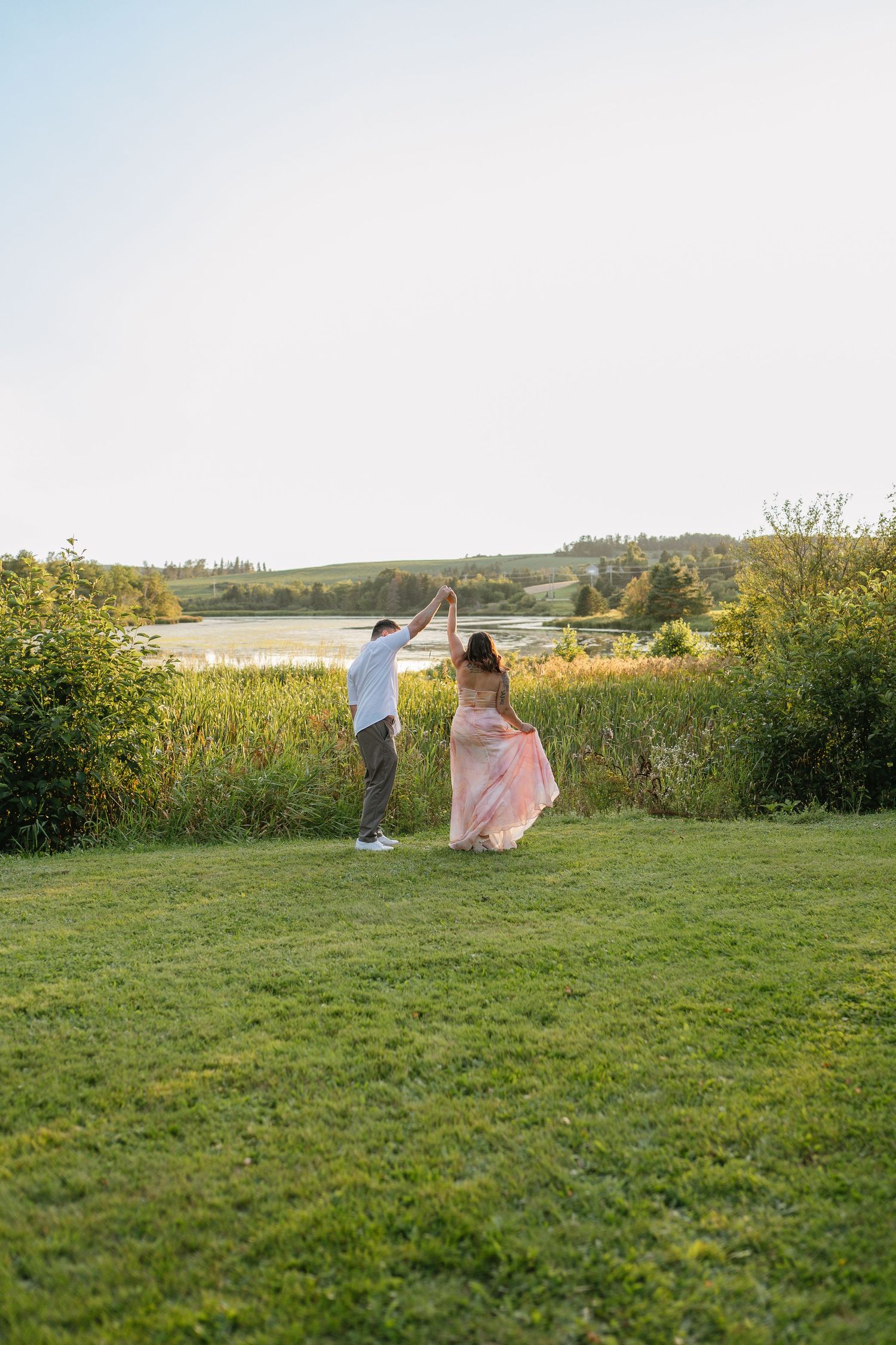 A couple dances outdoors on green grass near a lake, woman in flowing pink dress, man in white shirt, golden hour light.