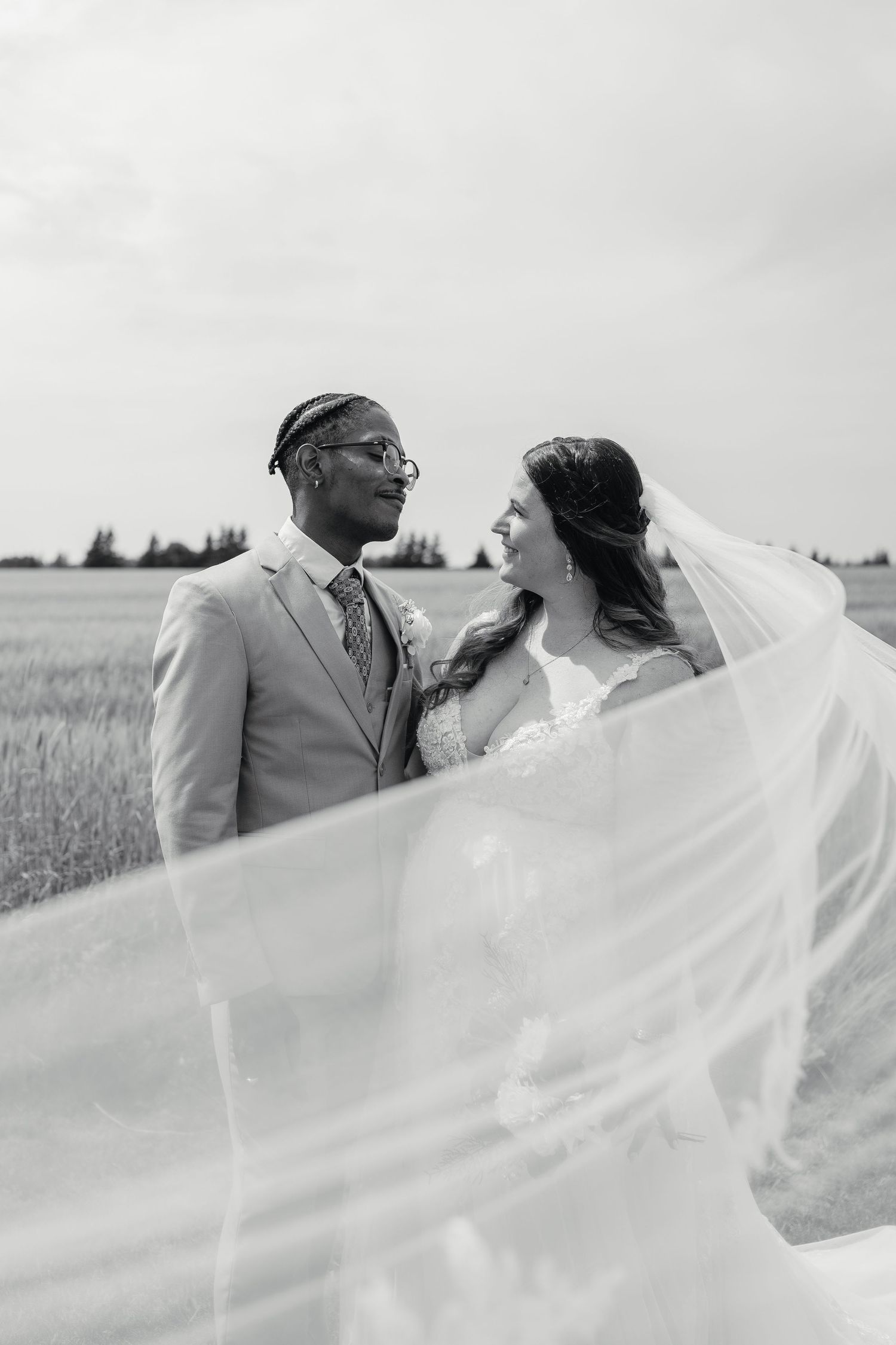Black and white portrait of interracial bride and groom gazing at each other as veil blows in the wind.