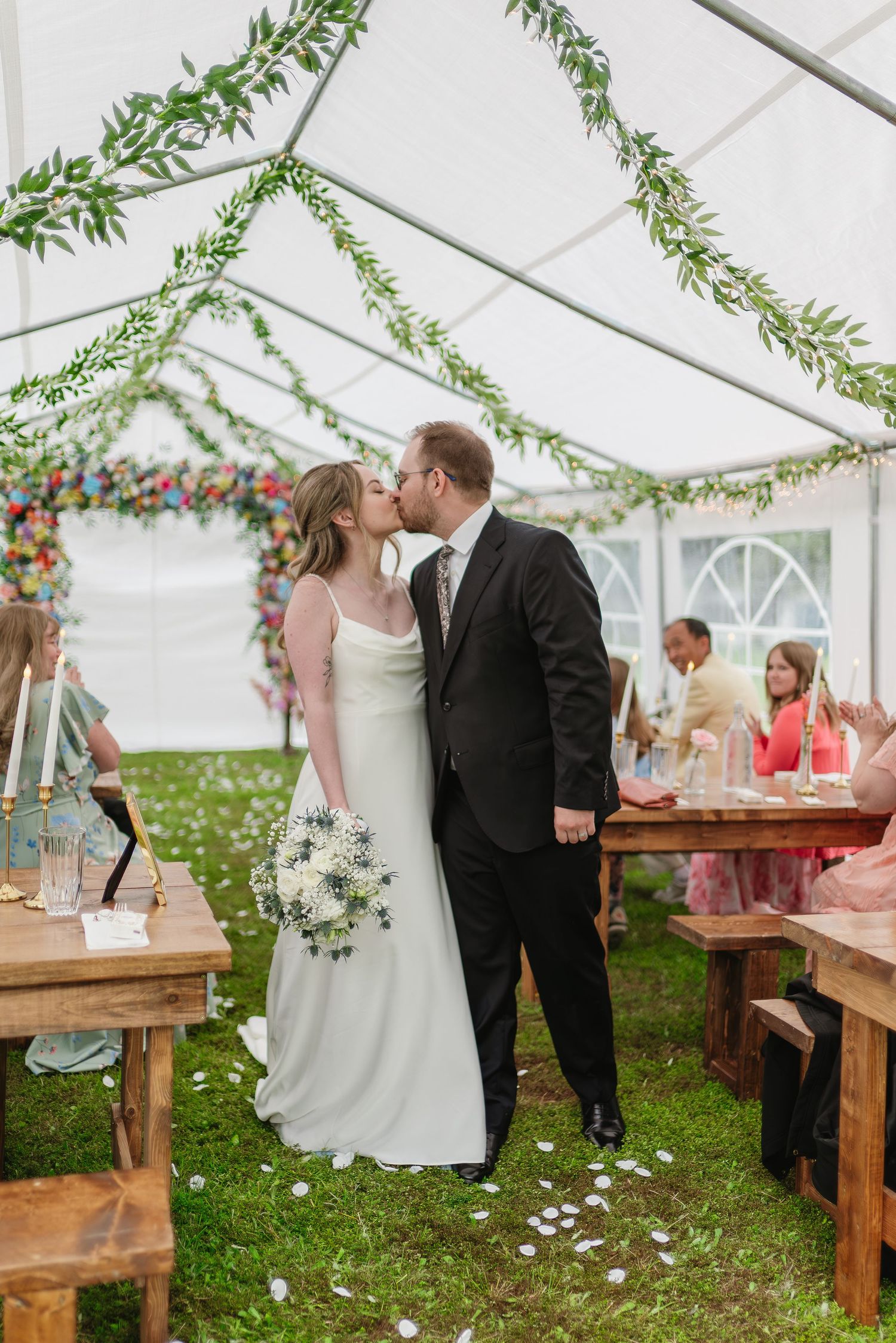 Newlyweds sharing first kiss as married couple in decorated marquee with guests seated nearby.