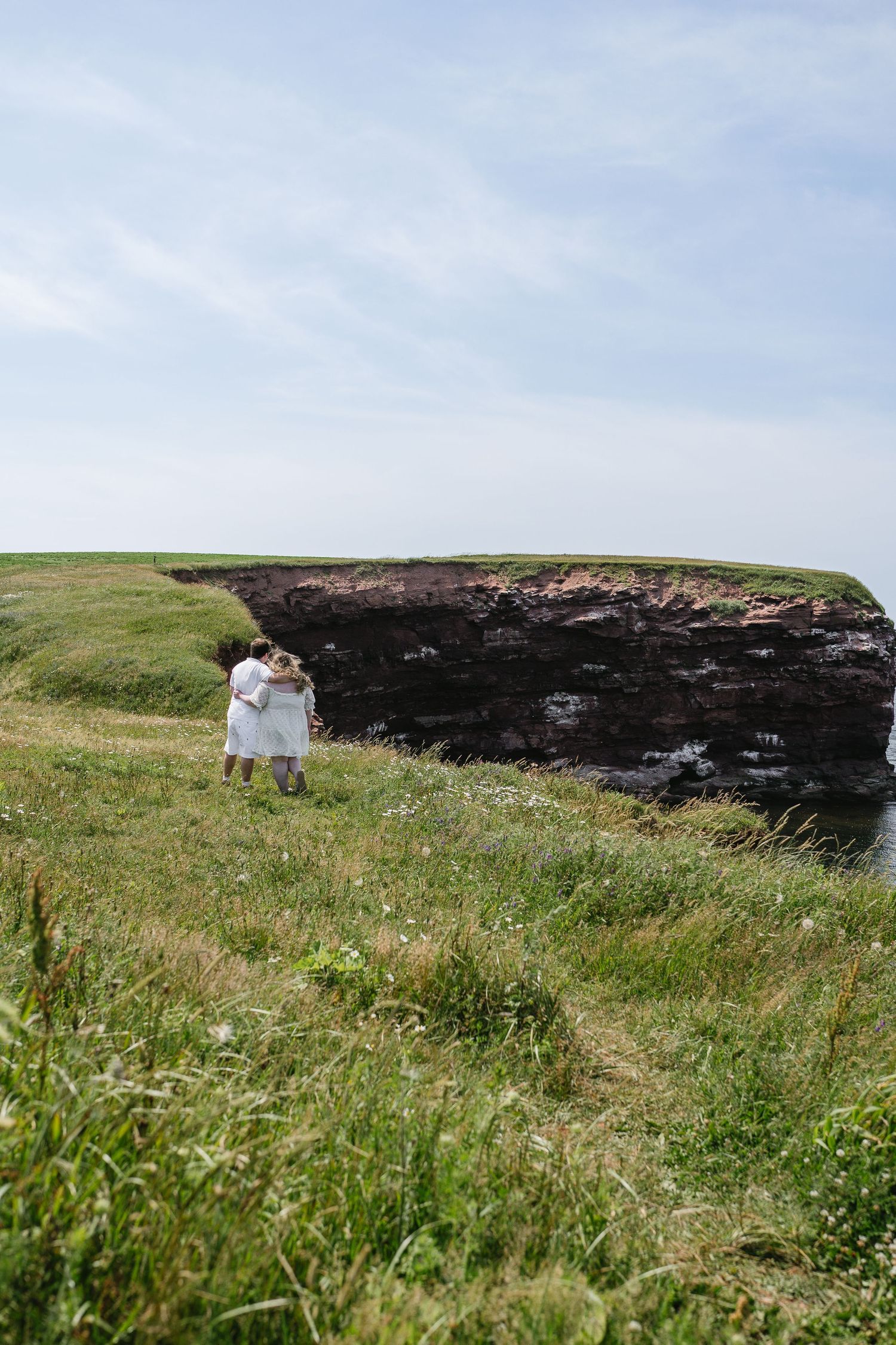 Couple in white walks arm in arm along a grassy coastal cliff edge overlooking the ocean on a clear day.