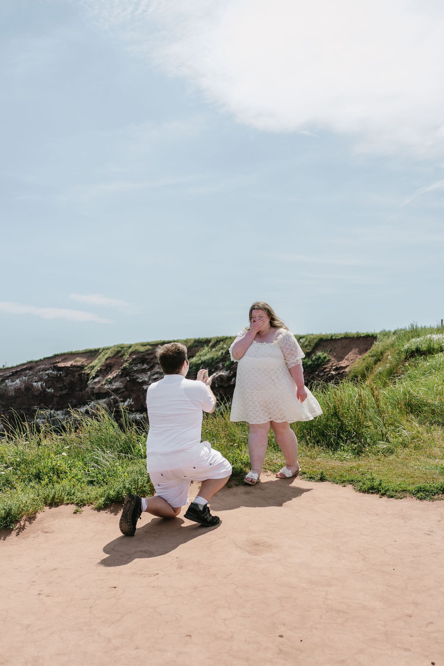 Emotional marriage proposal moment with man kneeling before tearful woman on coastal pathway.