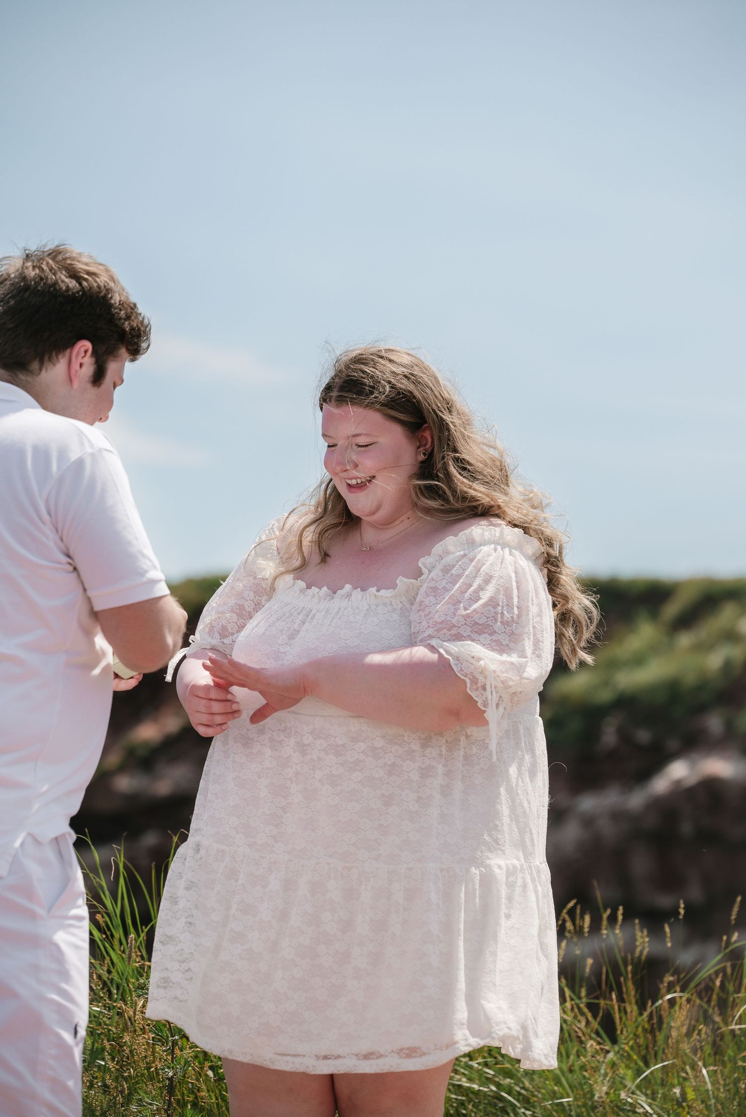 Newly engaged couple laughing together while admiring engagement ring on scenic coastal clifftop.