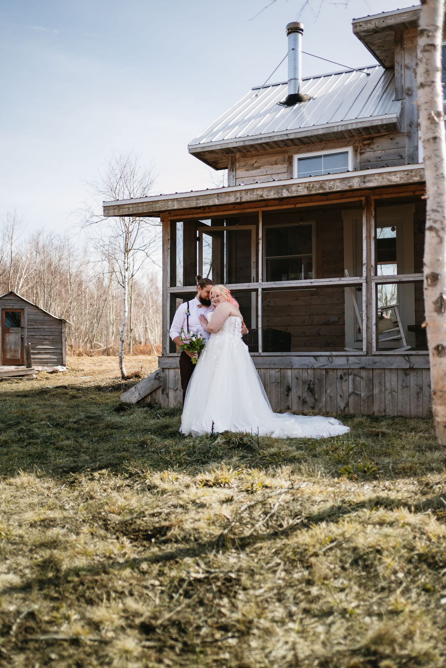 Bride in ballgown and groom embracing outside rustic wooden farmhouse during spring wedding.