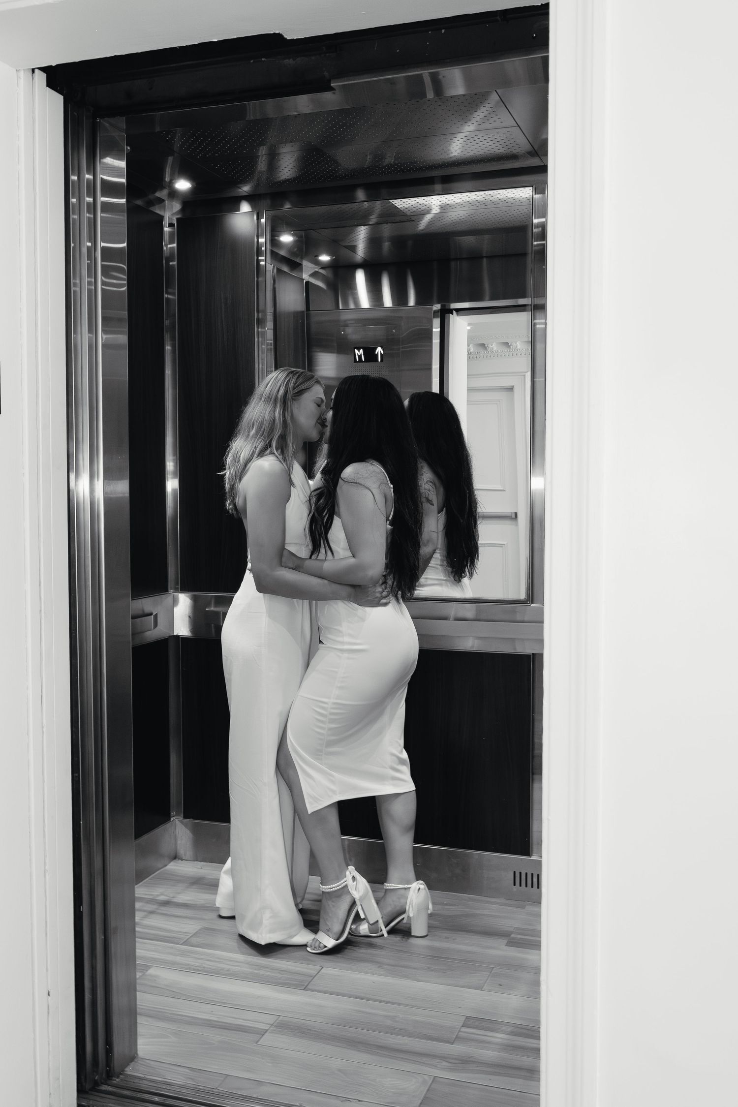 Black and white photo of two brides in white dresses sharing a kiss inside an elevator.
