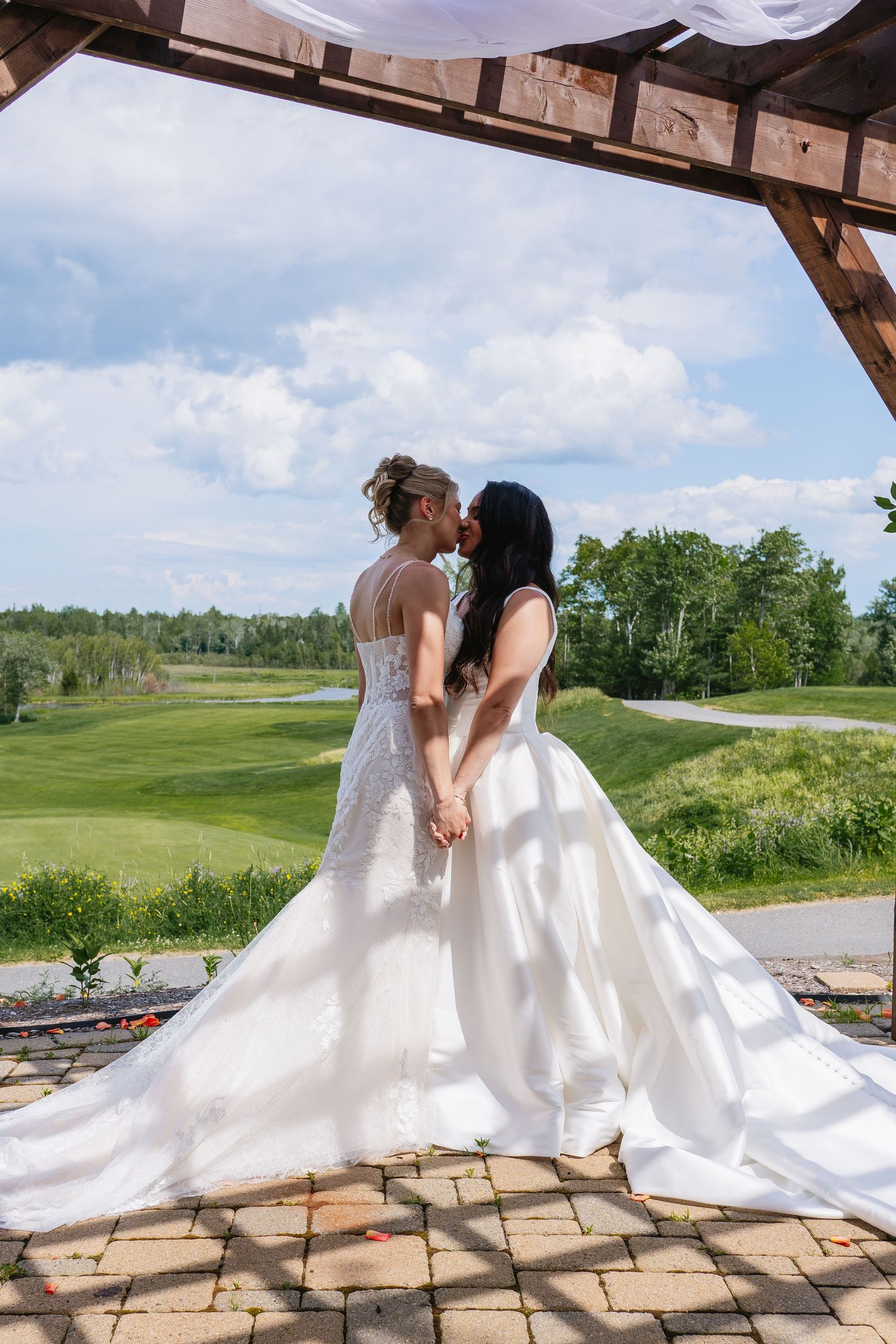Two brides share a romantic kiss under a wooden pergola overlooking a lush green golf course landscape.