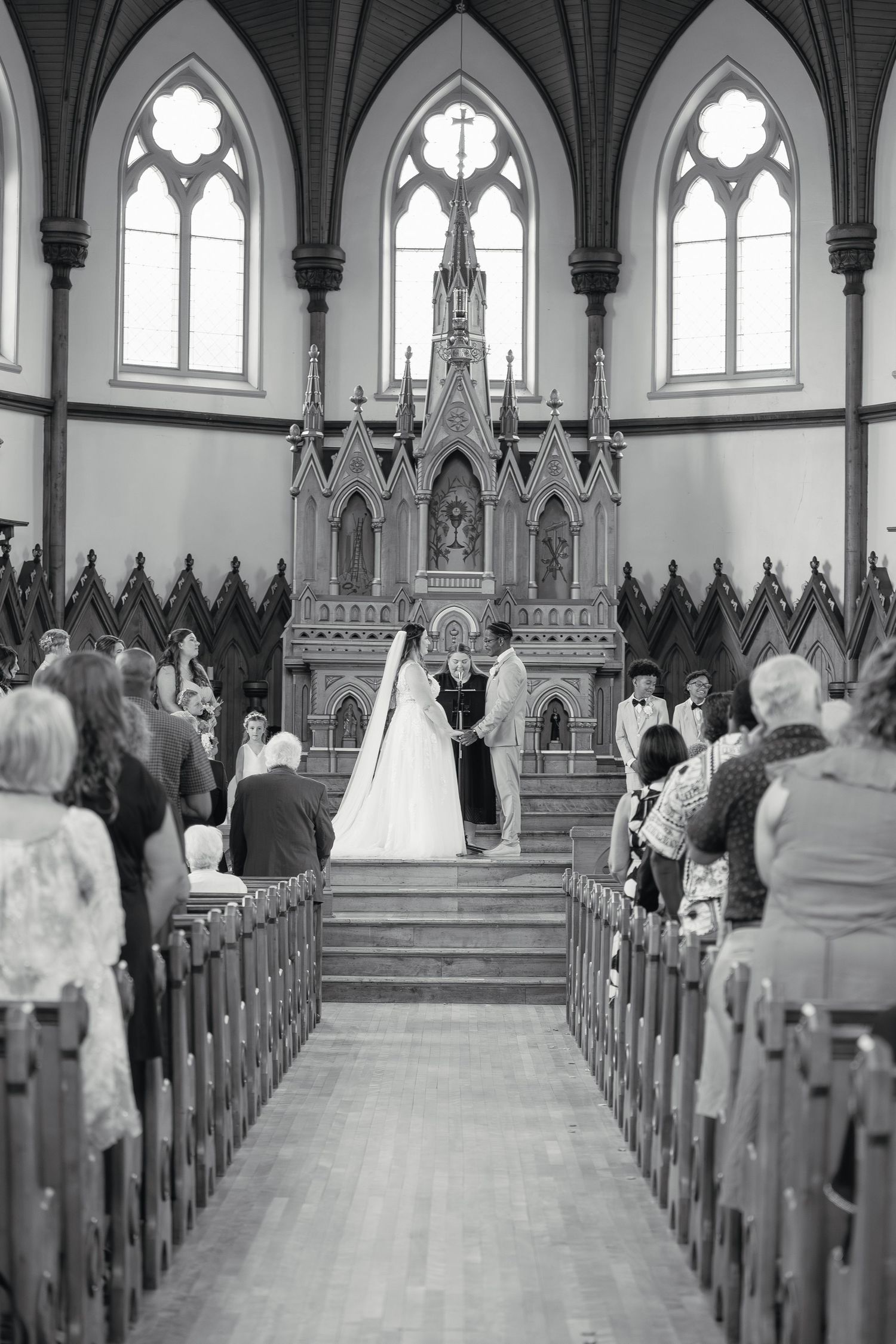 Black and white image of a wedding ceremony at a Gothic-style church altar with guests seated in wooden pews.