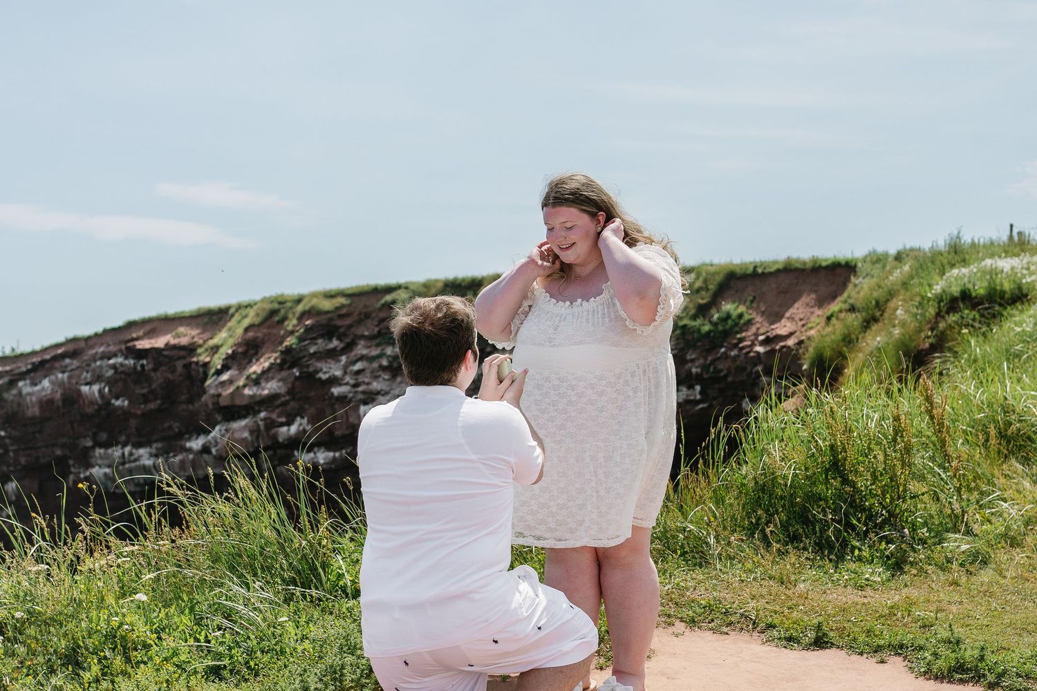 A man kneels to propose to an emotional woman in a white dress on a scenic coastal path.