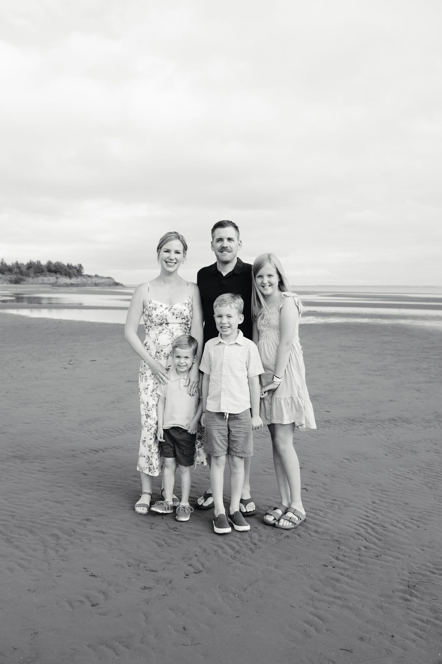 Black and white family portrait of five on a beach, two adults and three children smiling together on a cloudy day.