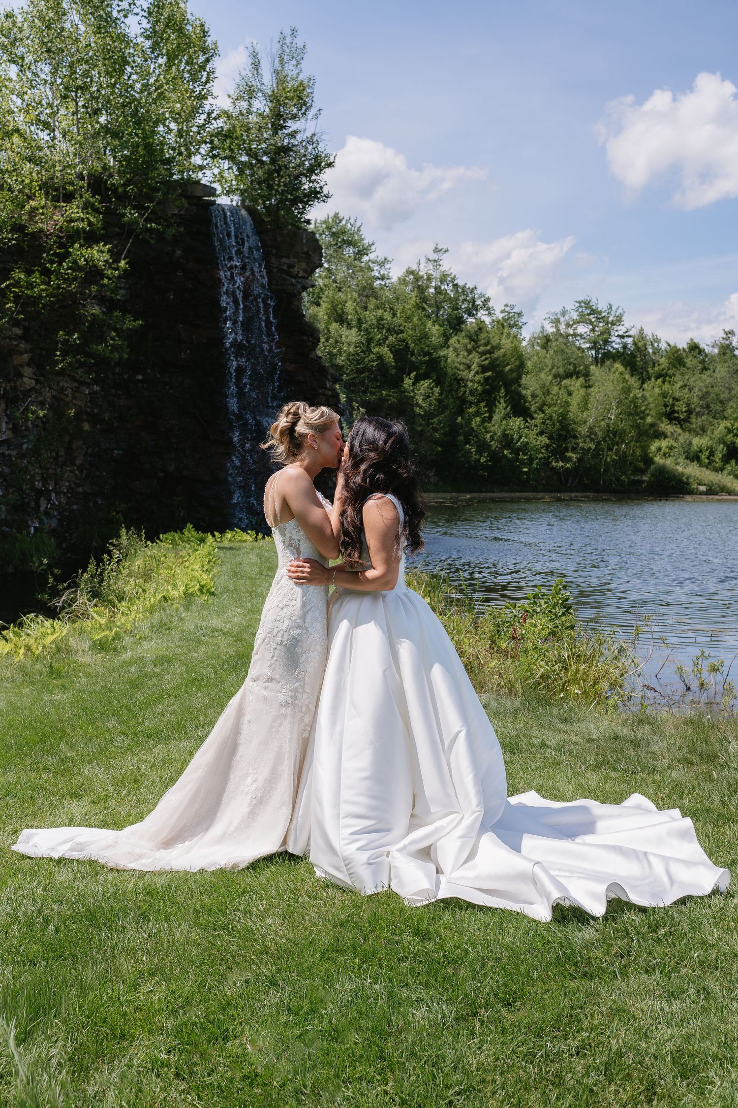 Two brides in elegant white gowns share a kiss near a scenic waterfall and lake on their wedding day.