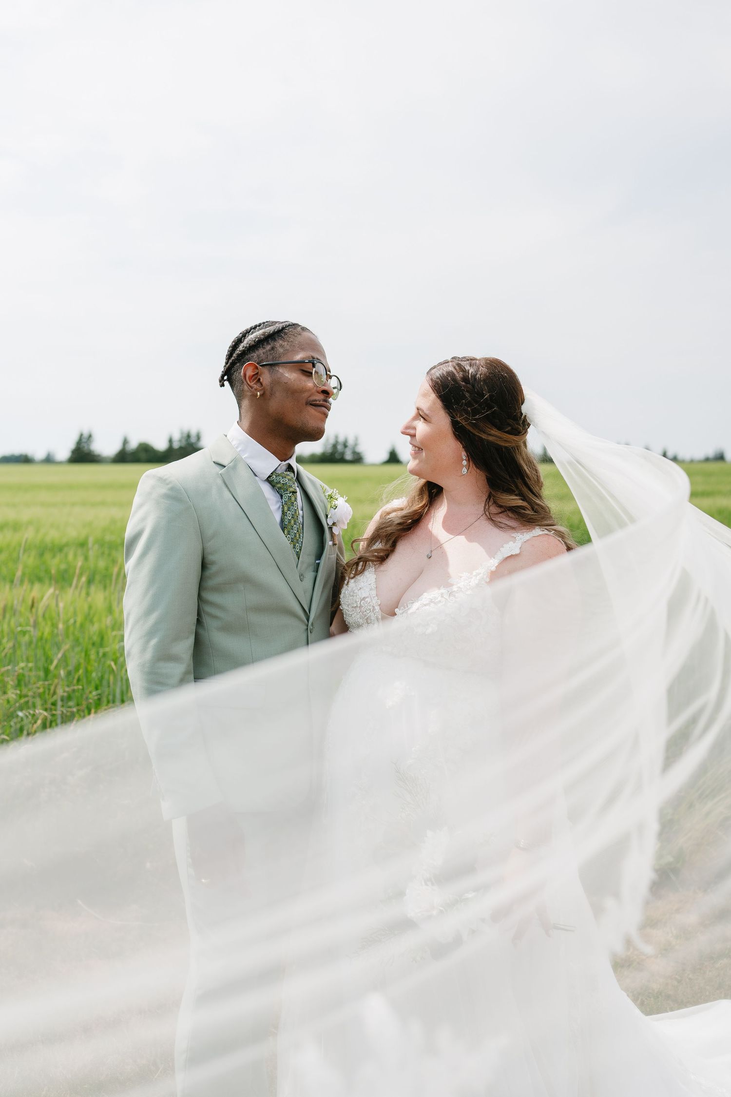 Bride and groom gazing at each other in a green field with the brides veil flowing in the wind.
