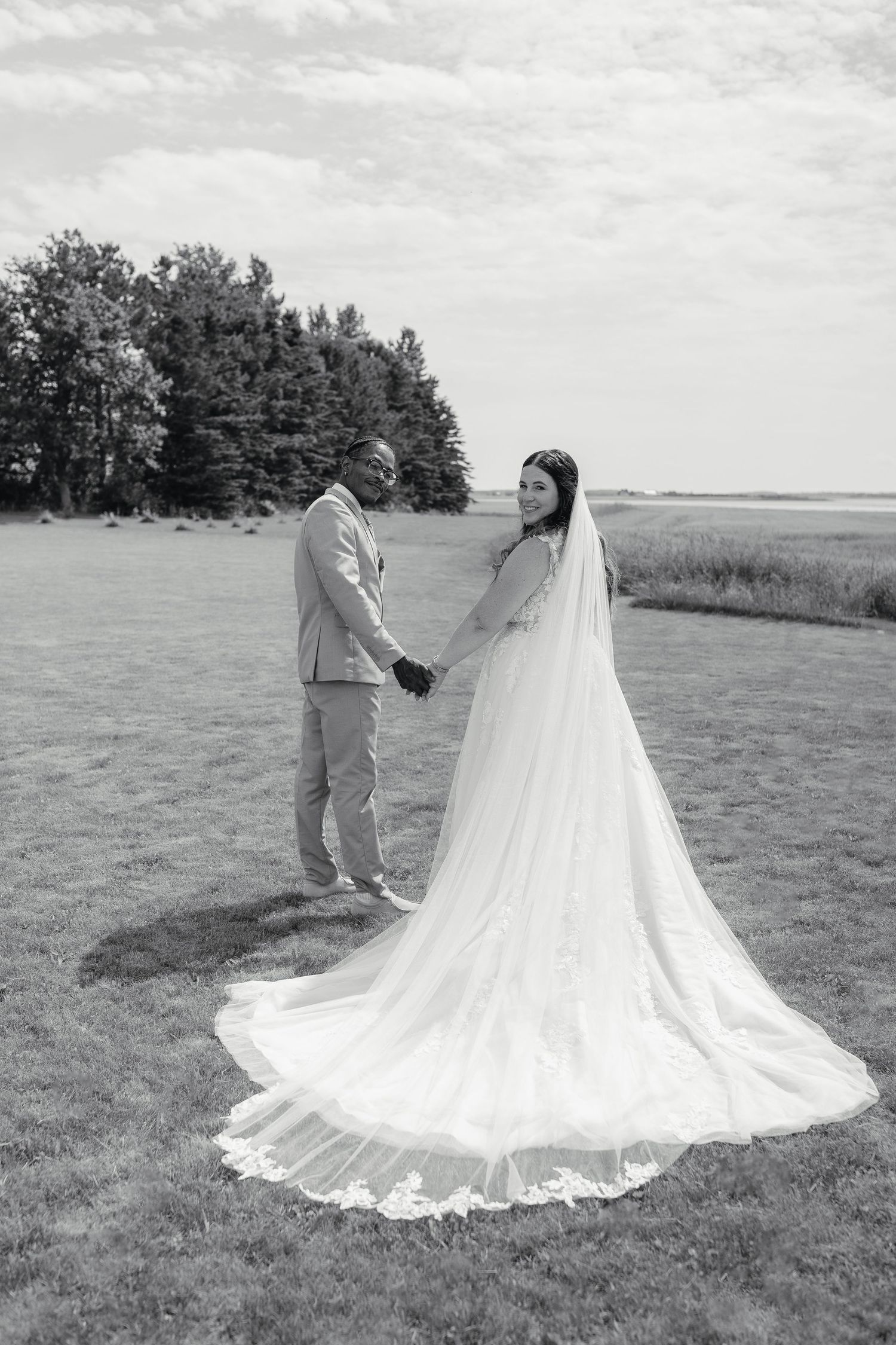 Black and white photo of bride and groom holding hands in an open field, bride wearing long cathedral veil.