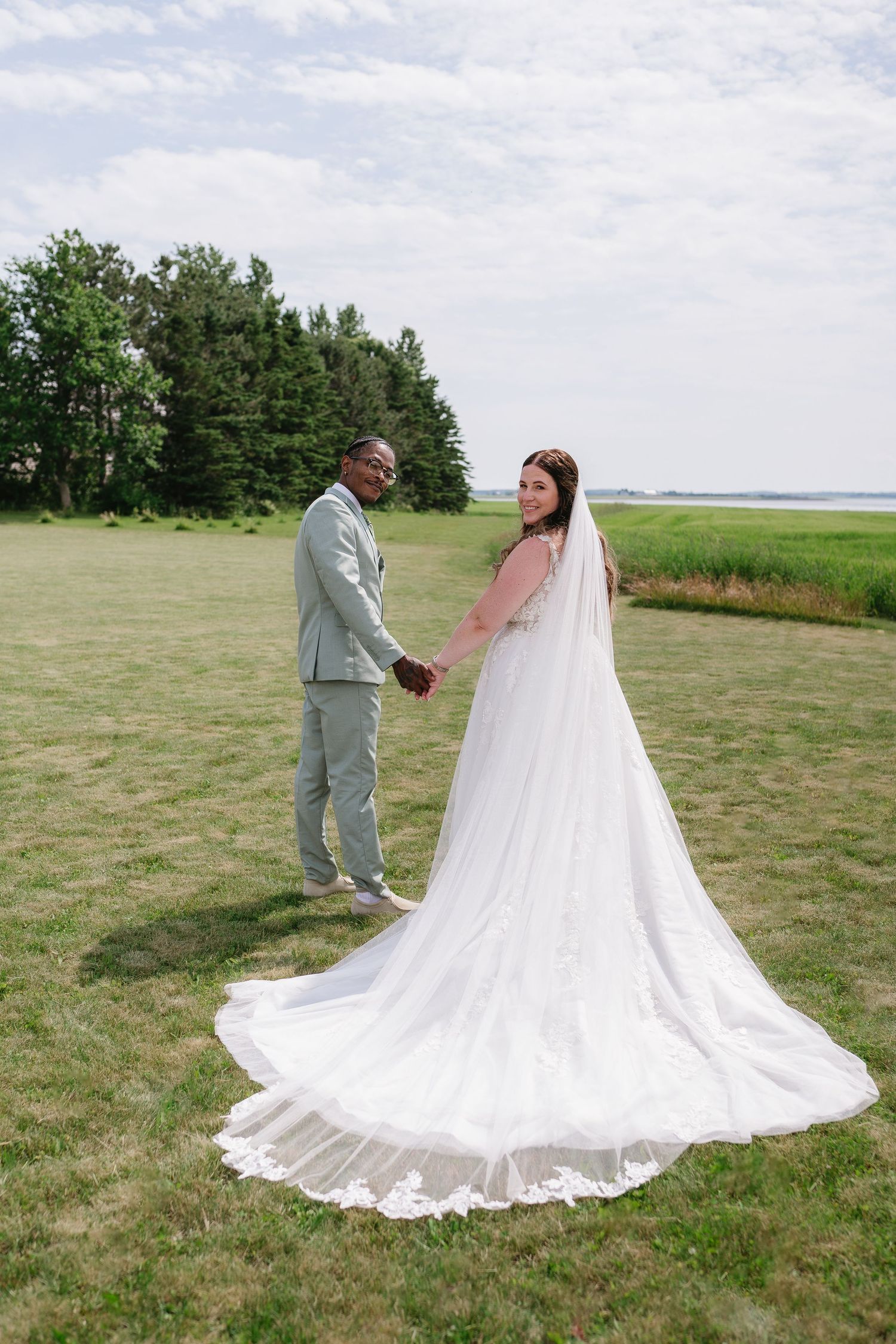 Interracial bride and groom hold hands in a green field, bride wearing a long lace cathedral veil.