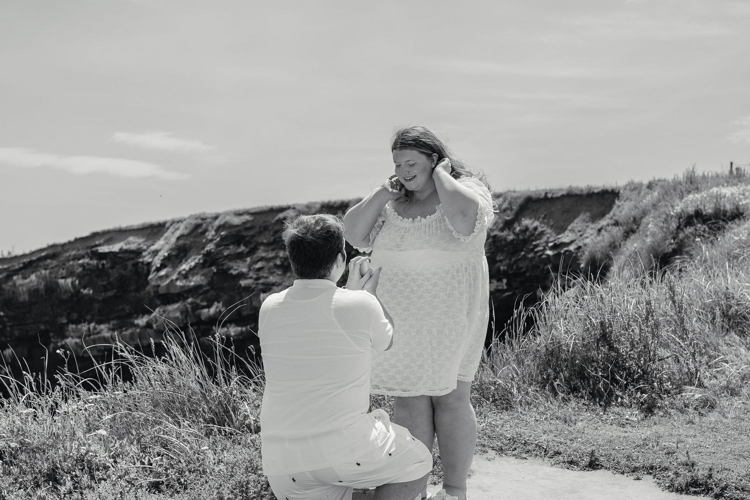 Black and white close-up of a man proposing on one knee to a surprised and emotional woman outdoors.