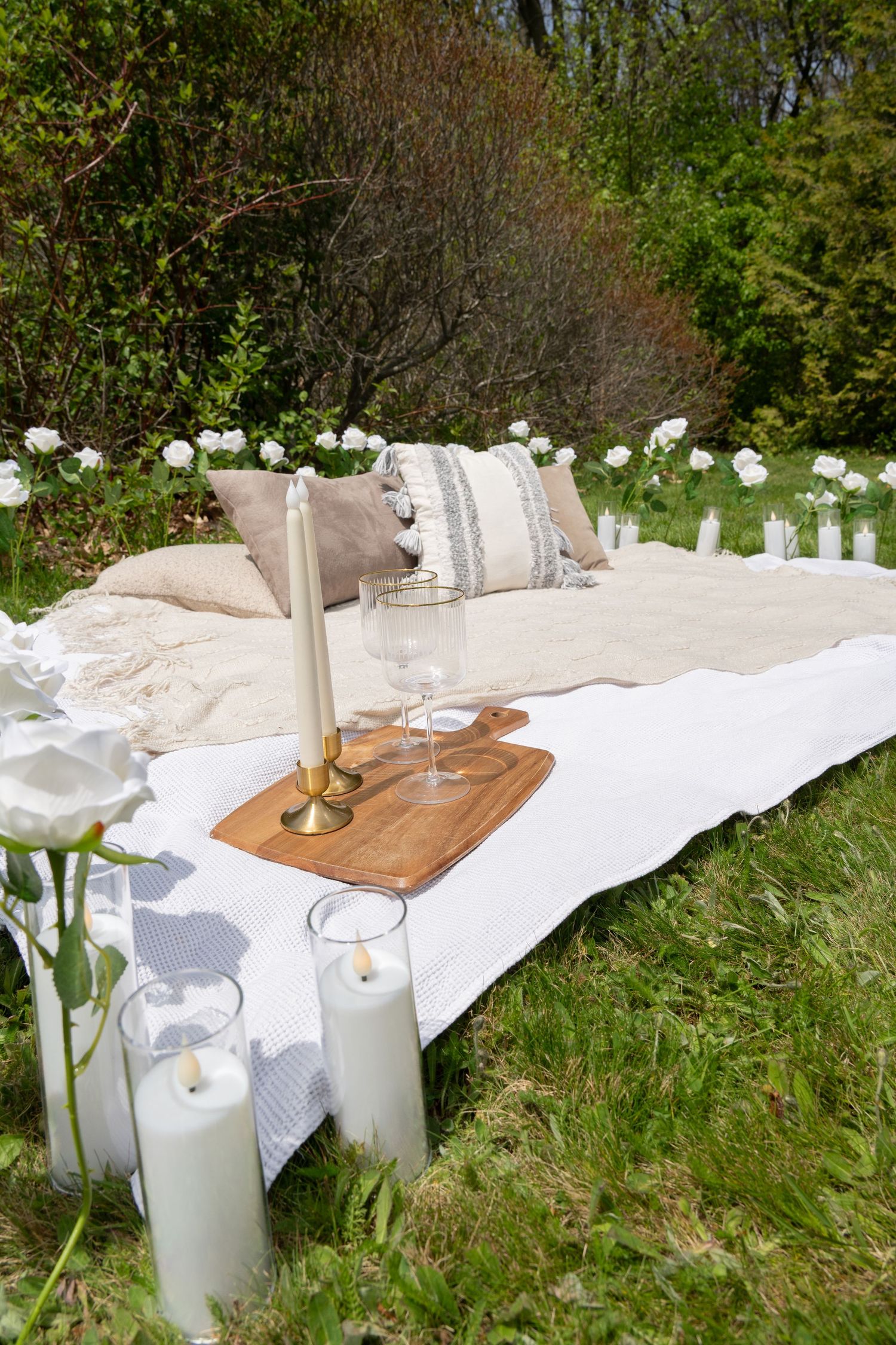 An elegant outdoor picnic setup with candles, wine glasses, and white roses on a white blanket.