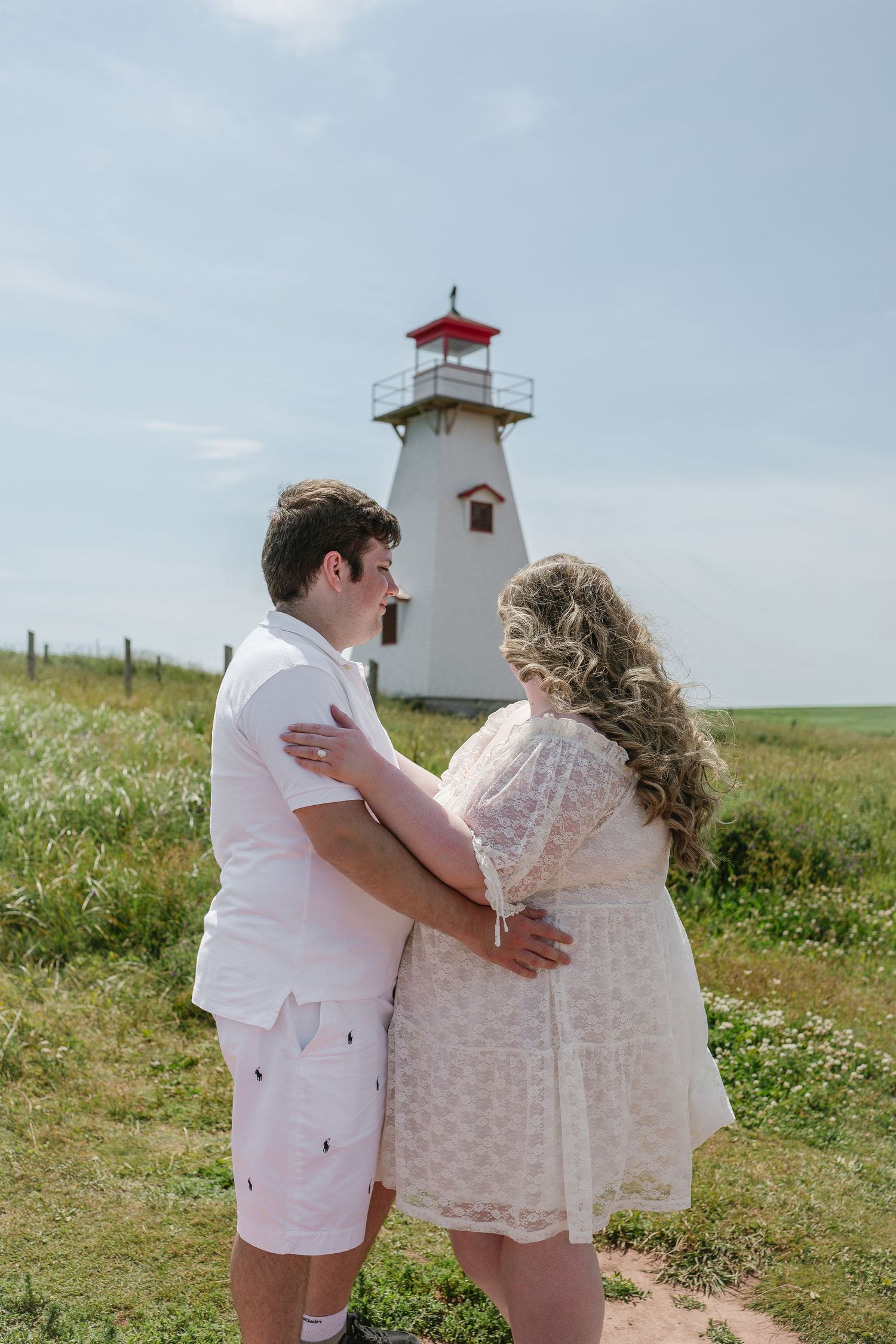 Couple in white outfits embracing near a red-topped lighthouse on a green coastal field under a blue sky.