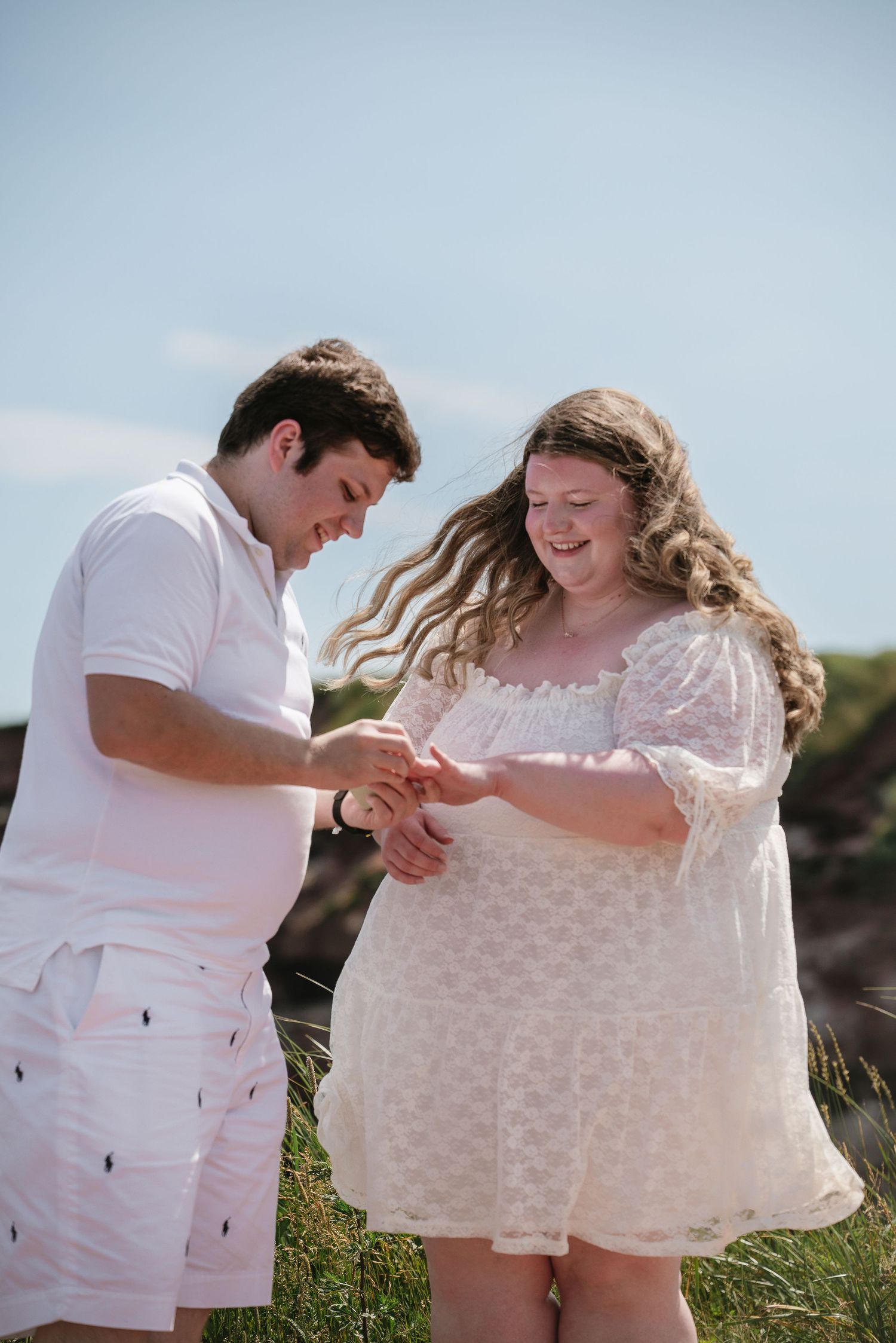 Happy couple smiling while examining engagement ring on grassy coastal clifftop on sunny day.