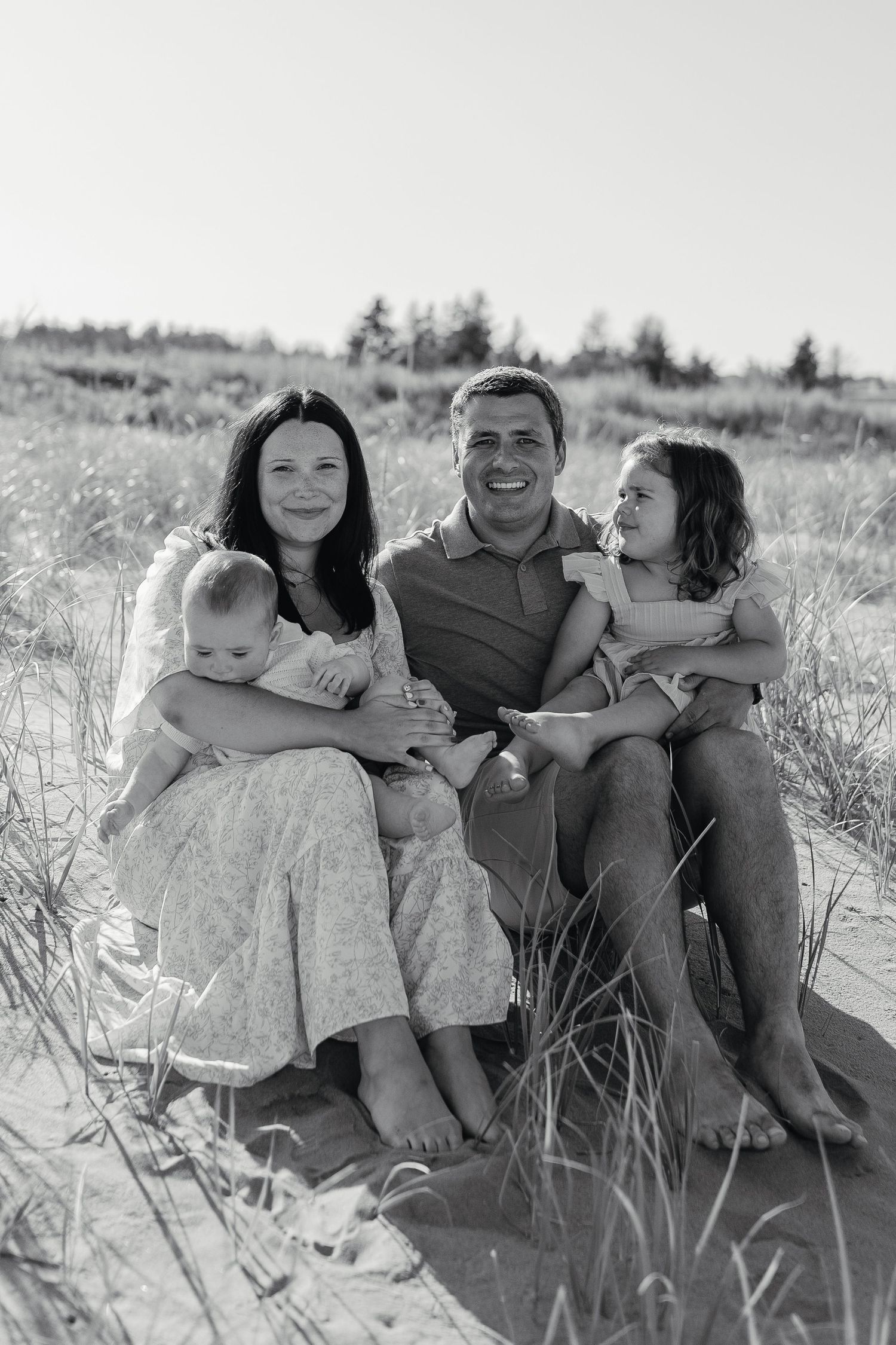Black and white family portrait on beach dunes with parents and two young children smiling.