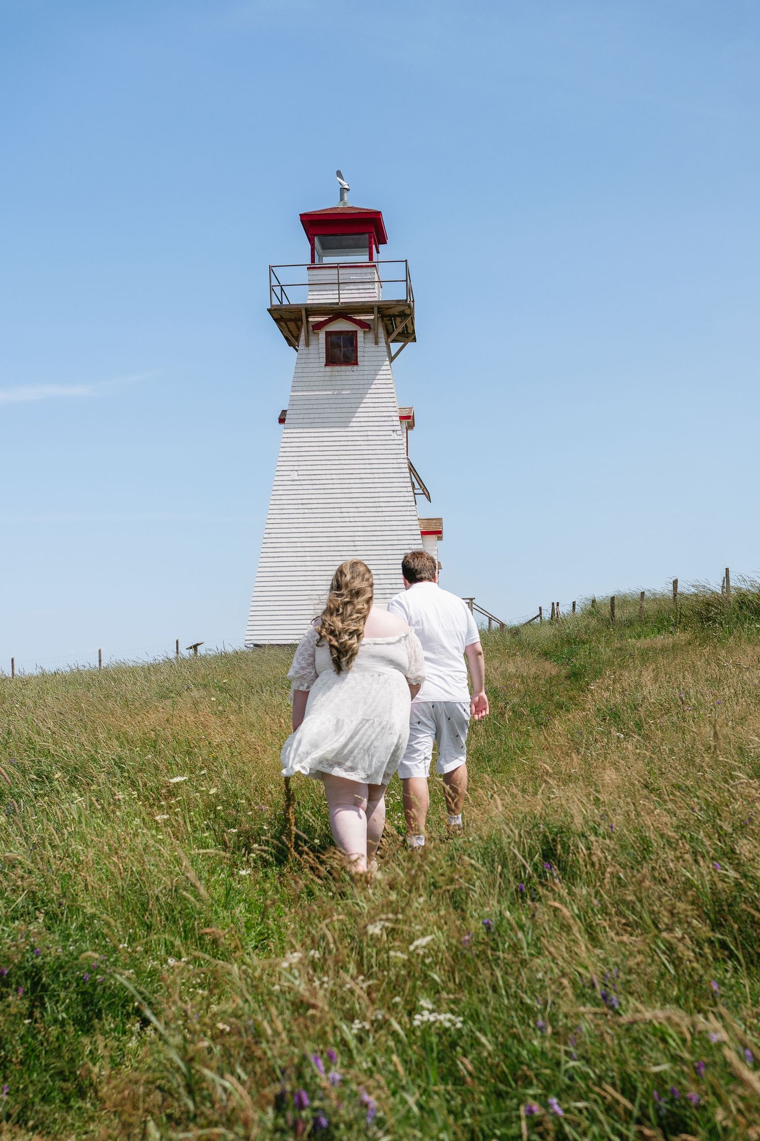 A couple in white walks toward a red and white lighthouse on a sunny day through a grassy field.