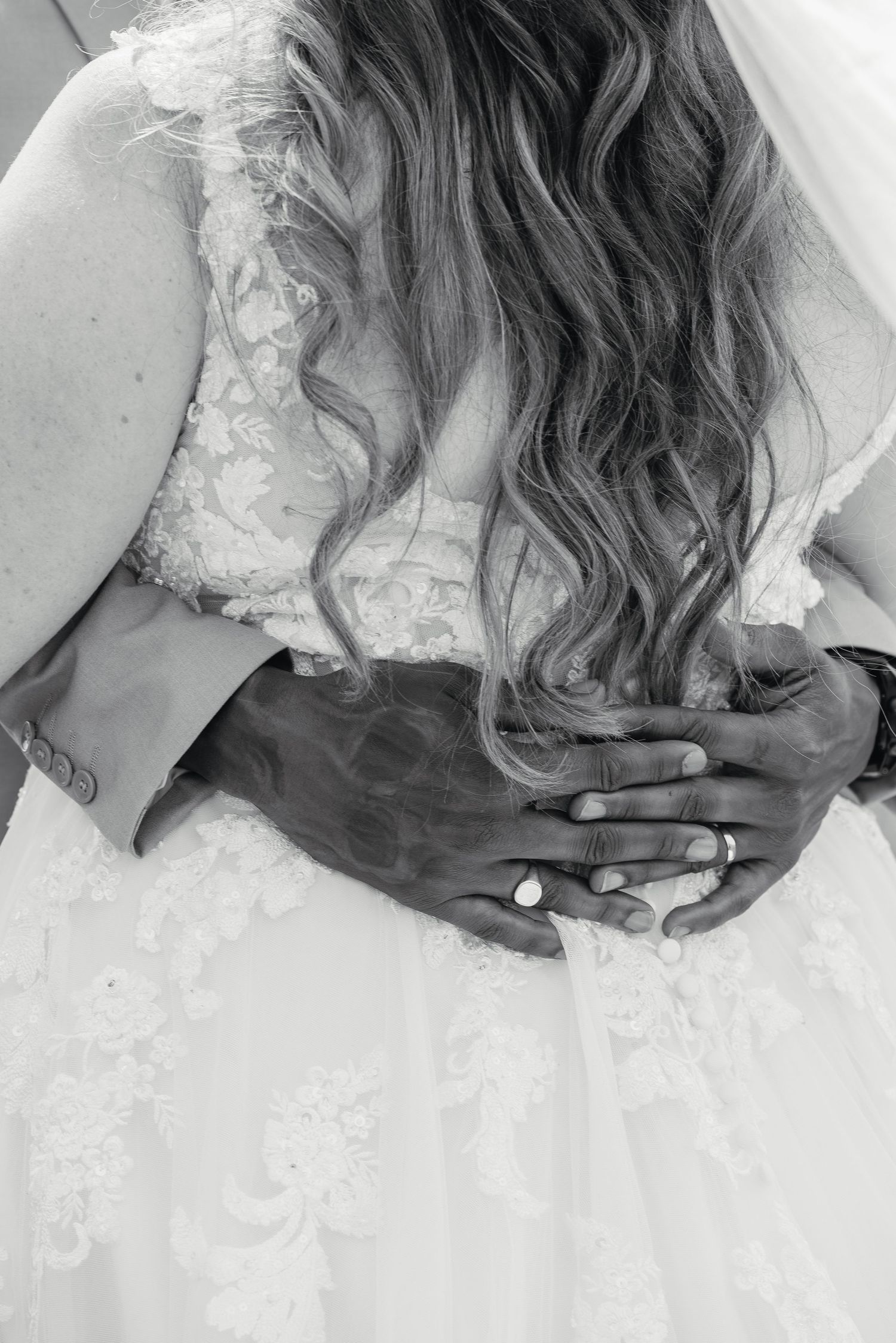 Close-up black and white photo of a groom's hands with wedding ring embracing his bride in a lace gown.