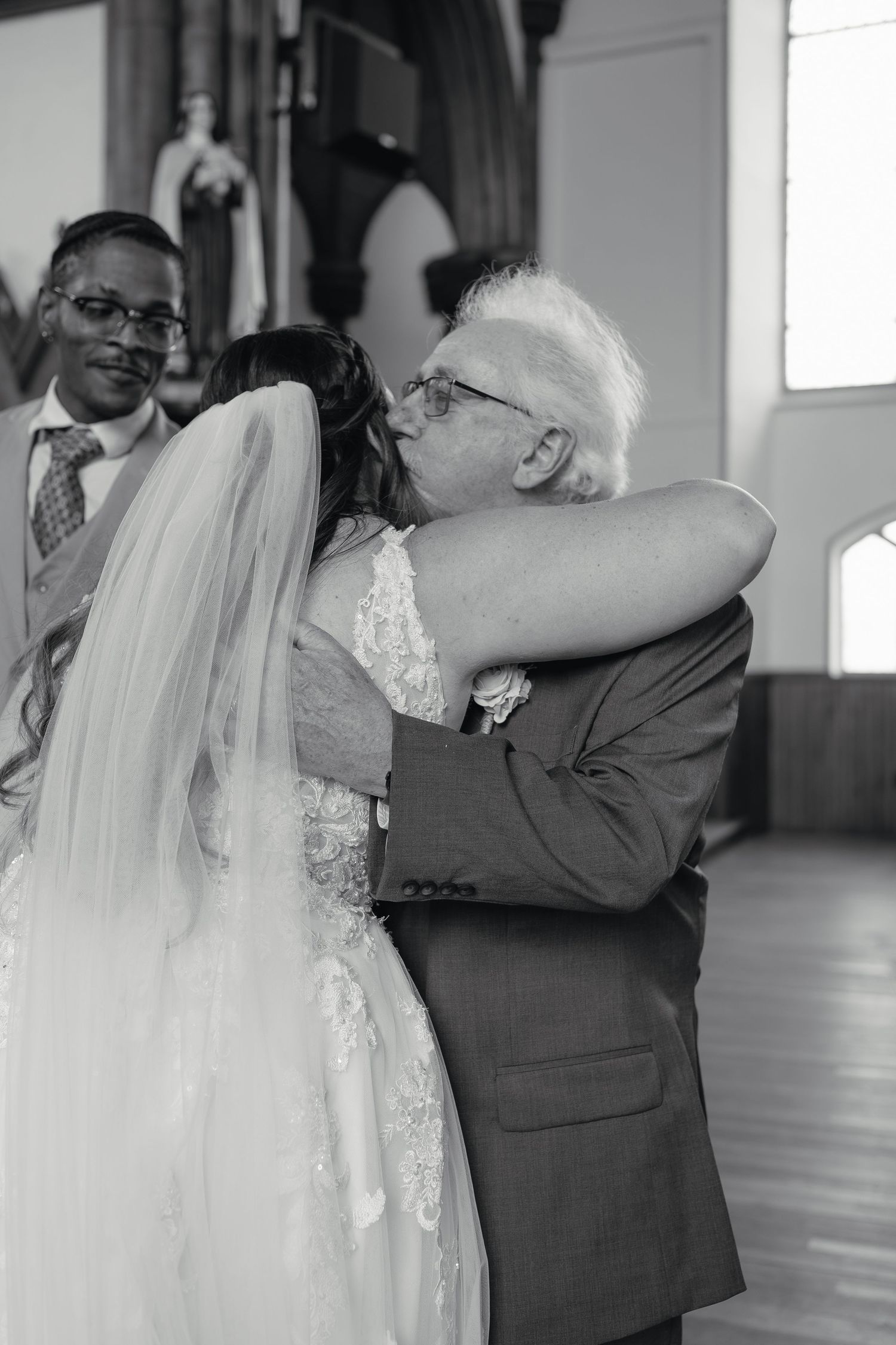 Black and white photo of a bride receiving an emotional hug from an older man during her wedding ceremony.