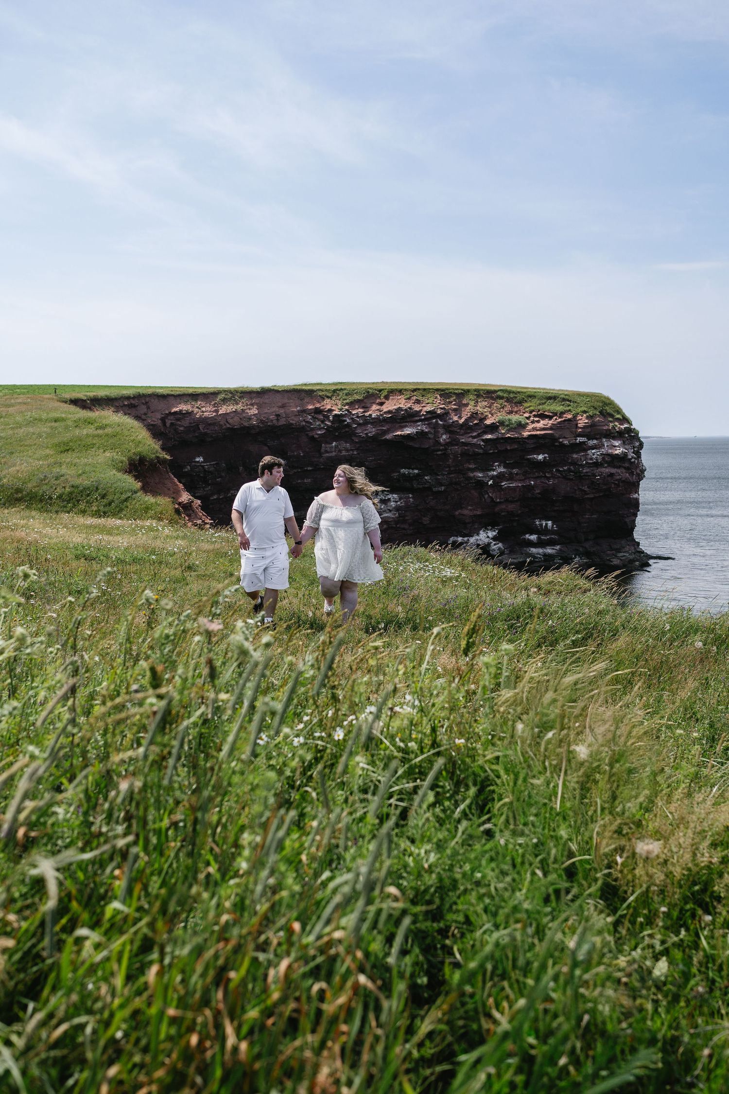 A couple in white outfits walks hand in hand through green coastal cliffs overlooking the ocean.