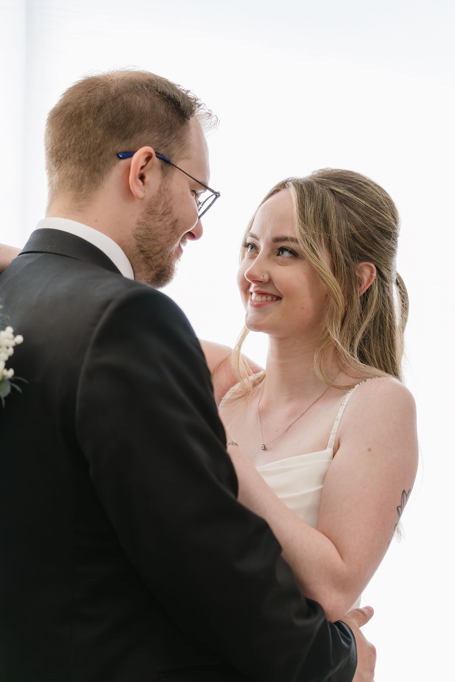 Smiling blonde bride in white dress embraces glasses-wearing groom in black suit indoors.