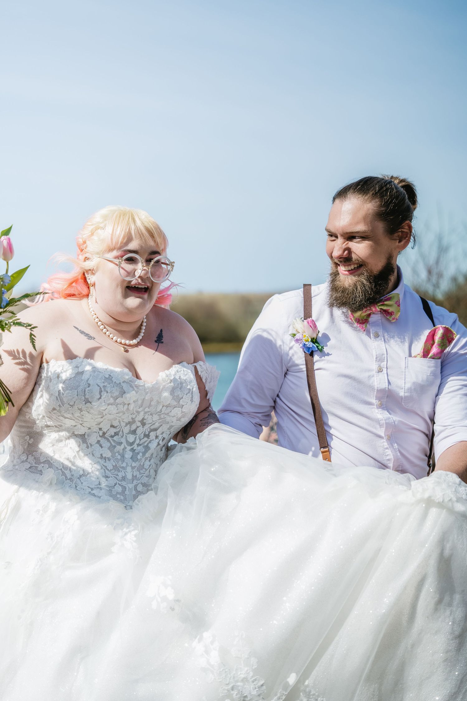 Smiling bride in lace gown and bearded groom in suspenders laughing outdoors by a lake in color photo.