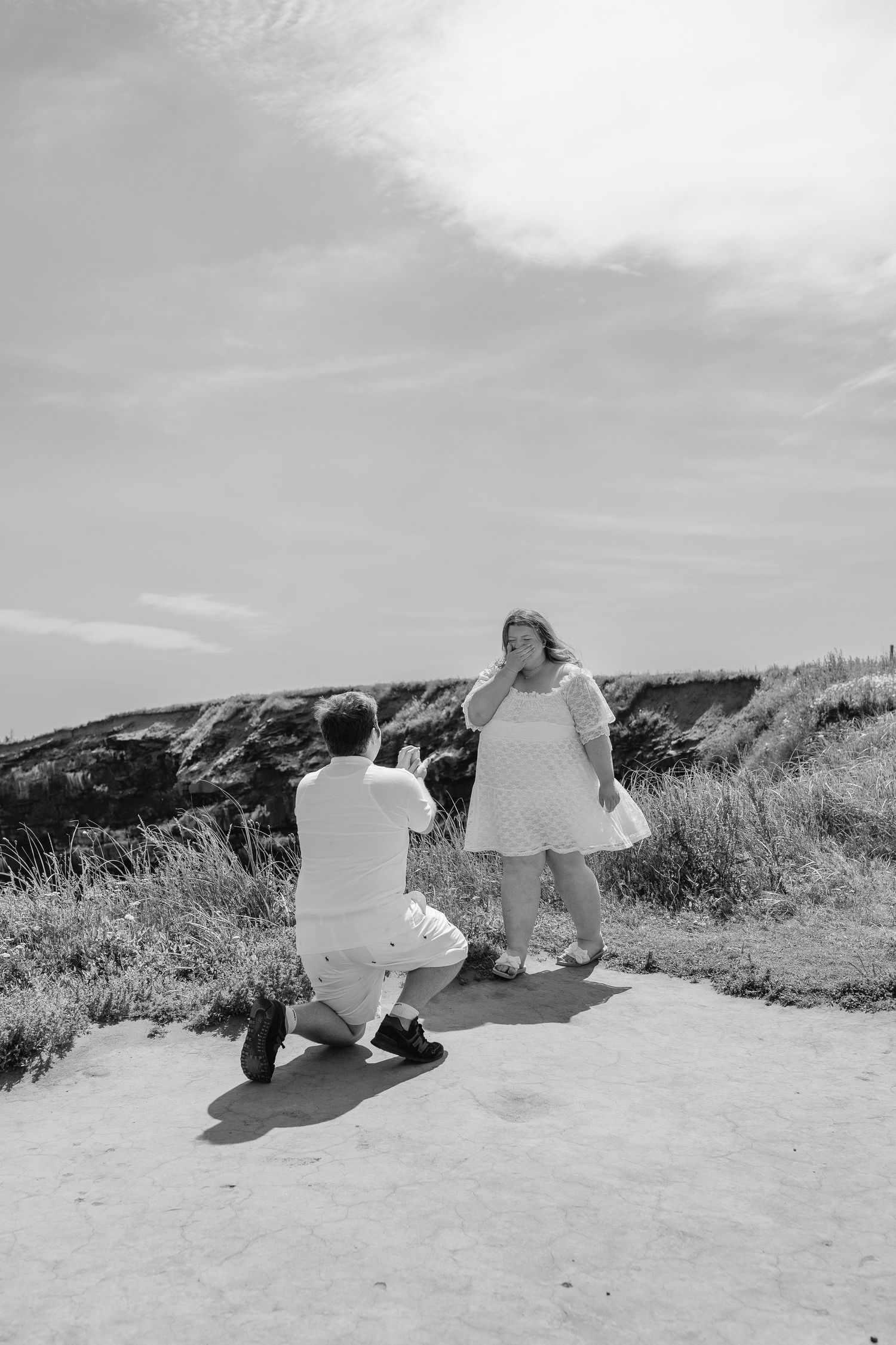 Man proposing on one knee to emotional woman on scenic outdoor path with grassy hills and cloudy sky.