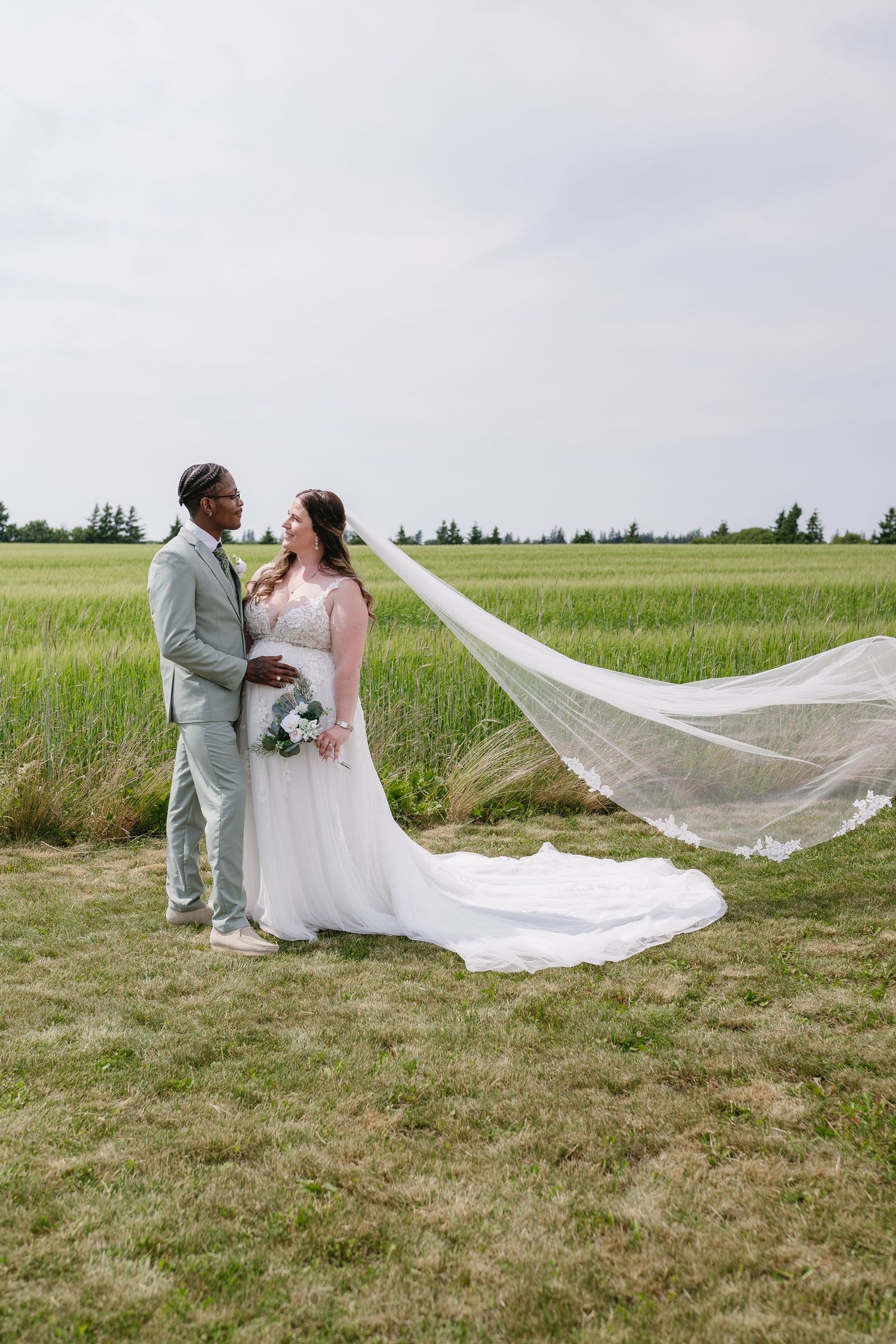 Interracial bride and groom stand in a green field as the bride's cathedral veil dramatically billows in the wind.