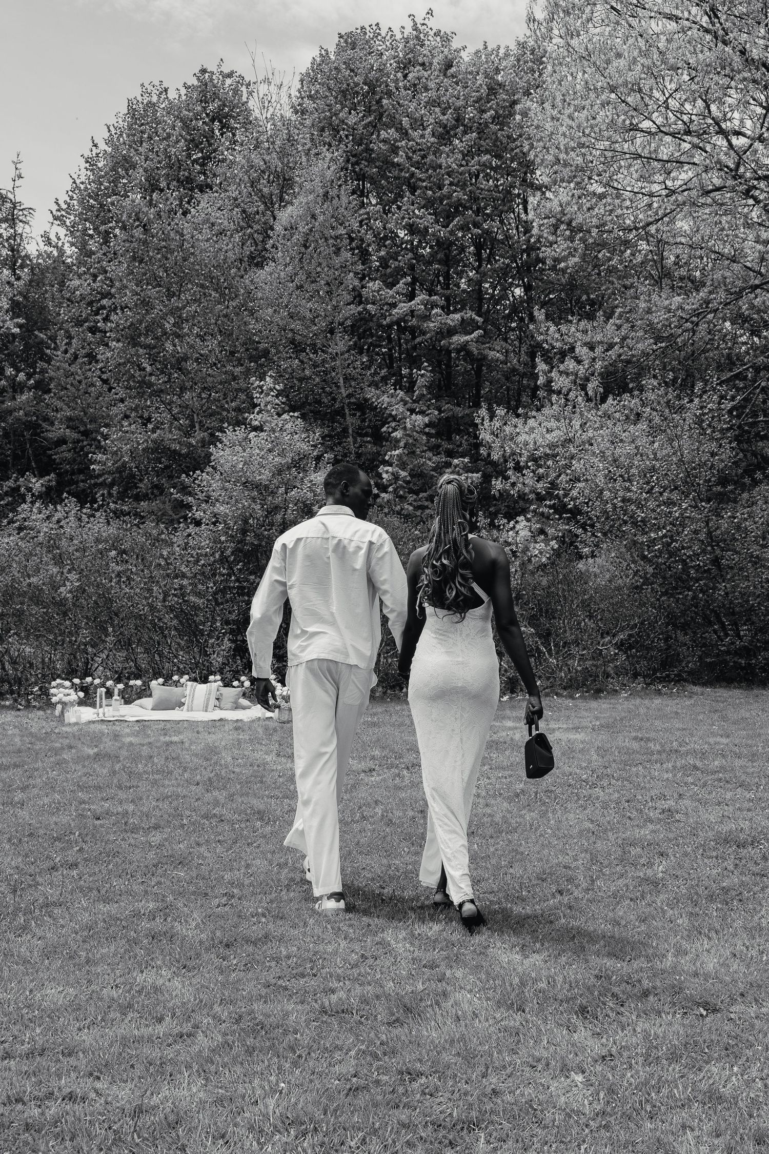 Black and white photo of a couple in white attire walking hand in hand across a lush green lawn toward trees.