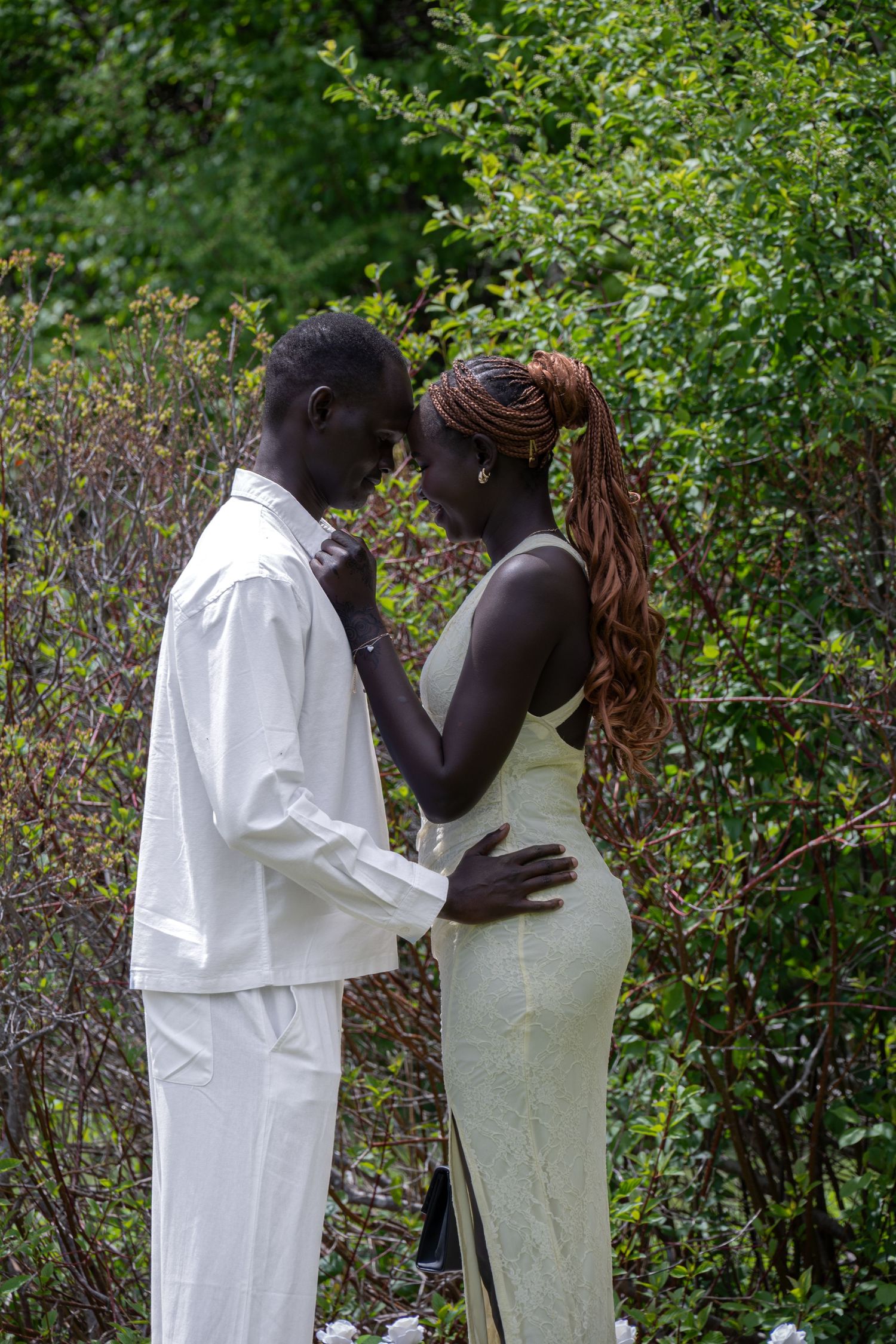 Couple sharing an intimate forehead-to-forehead moment surrounded by lush green bushes in a garden.