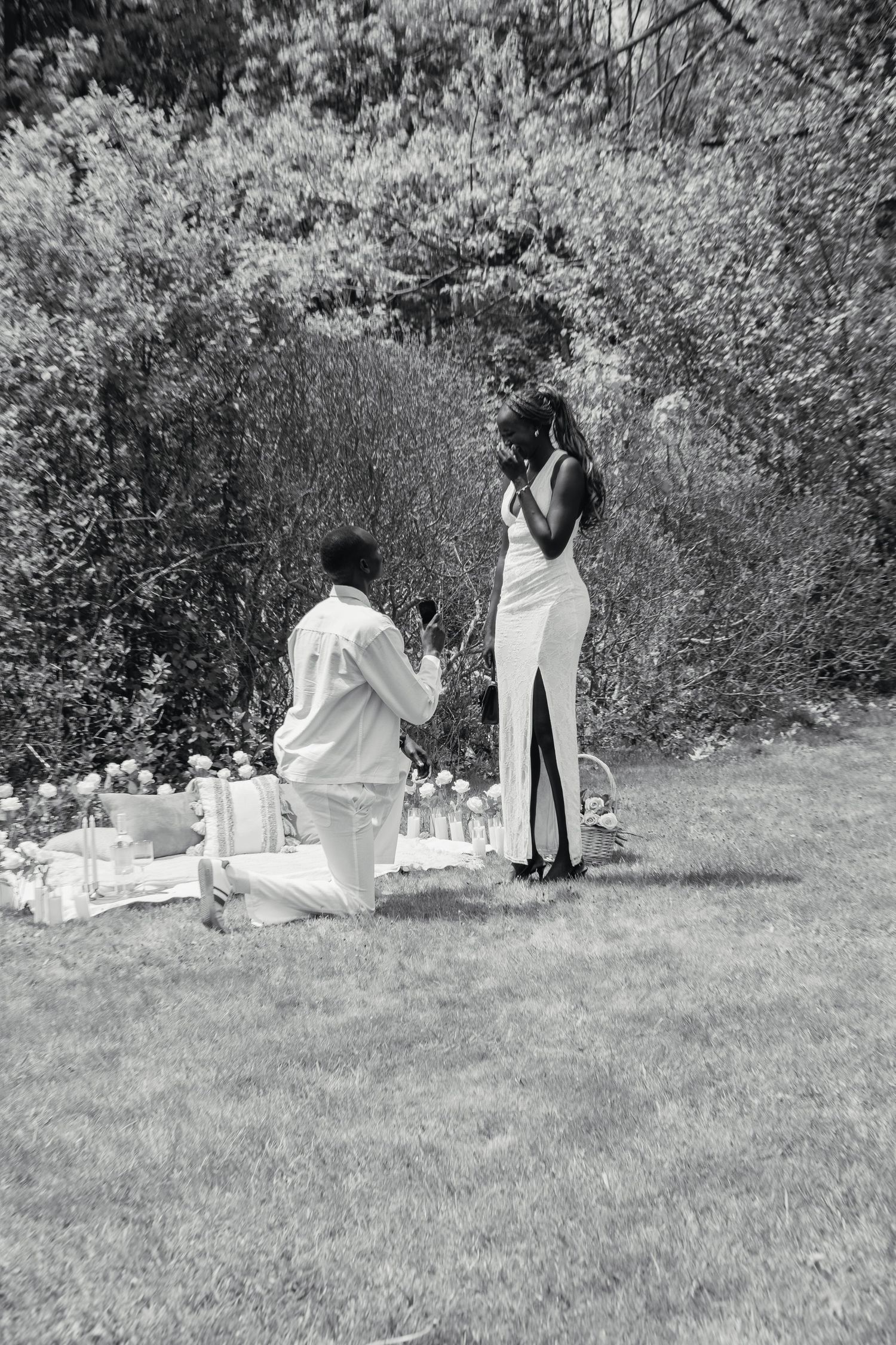 Black and white photo of a man on one knee proposing to a woman in a white dress in a garden setting.