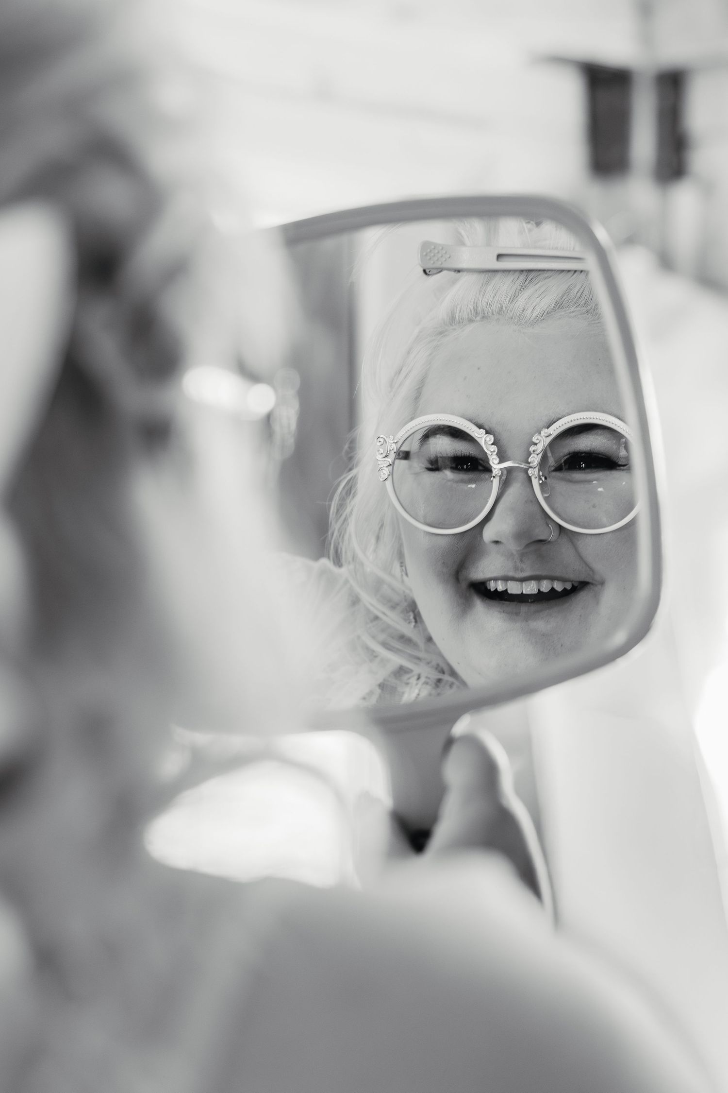 Bride smiling at her reflection in a handheld mirror wearing decorative round glasses, black and white photo.