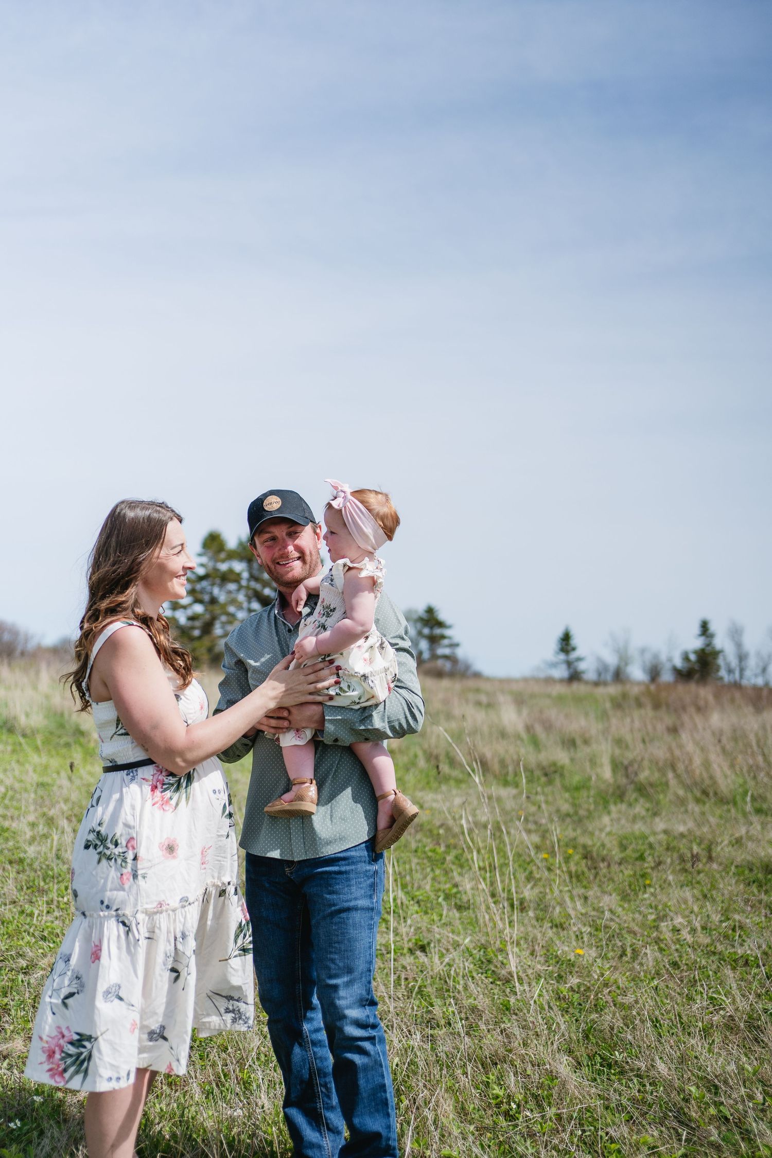 Smiling parents holding their baby girl in a bright sunny meadow with blue sky above.