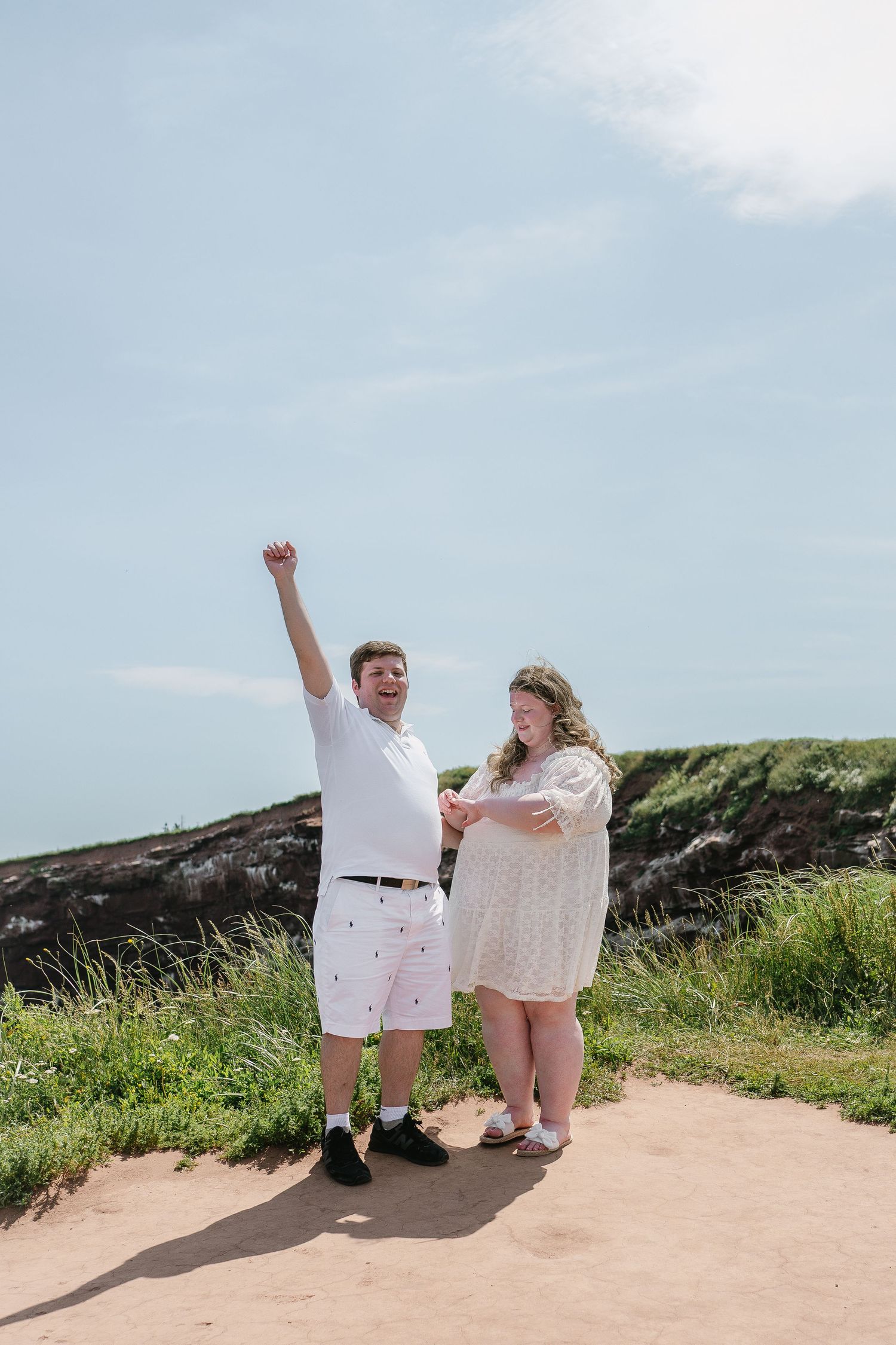 Excited newly engaged couple celebrating with raised fist and joyful expressions on coastal path.