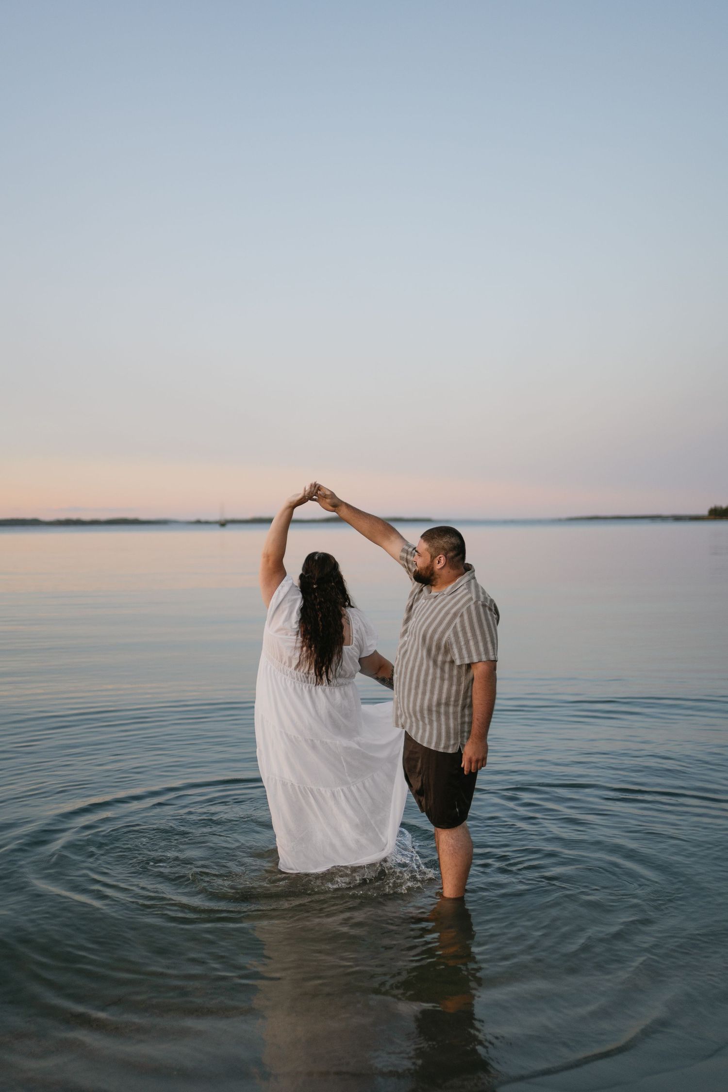 Couple dances together standing in calm ocean water at sunset, woman in flowing white dress being twirled.