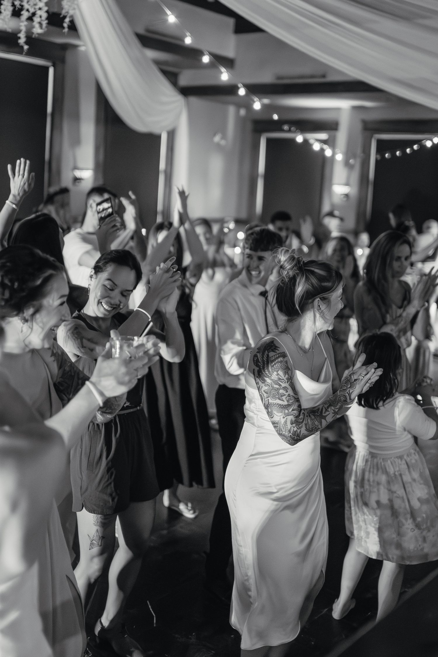 Black and white photo of two brides and wedding guests dancing joyfully on the reception dance floor.