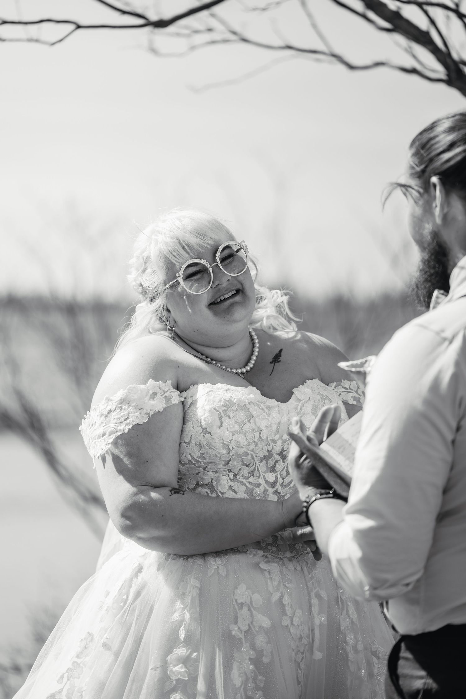 Bride in lace off-shoulder wedding gown laughing joyfully during outdoor wedding ceremony, black and white.
