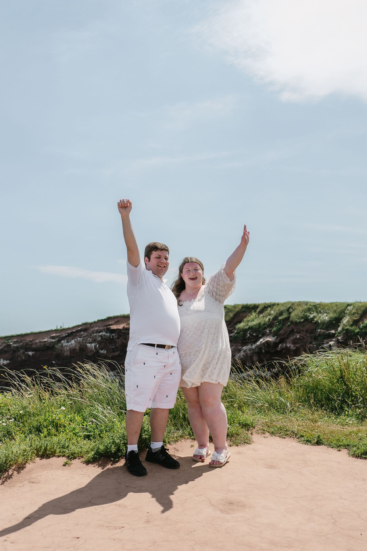 Jubilant newly engaged couple raising arms in celebration on a sunny coastal clifftop.