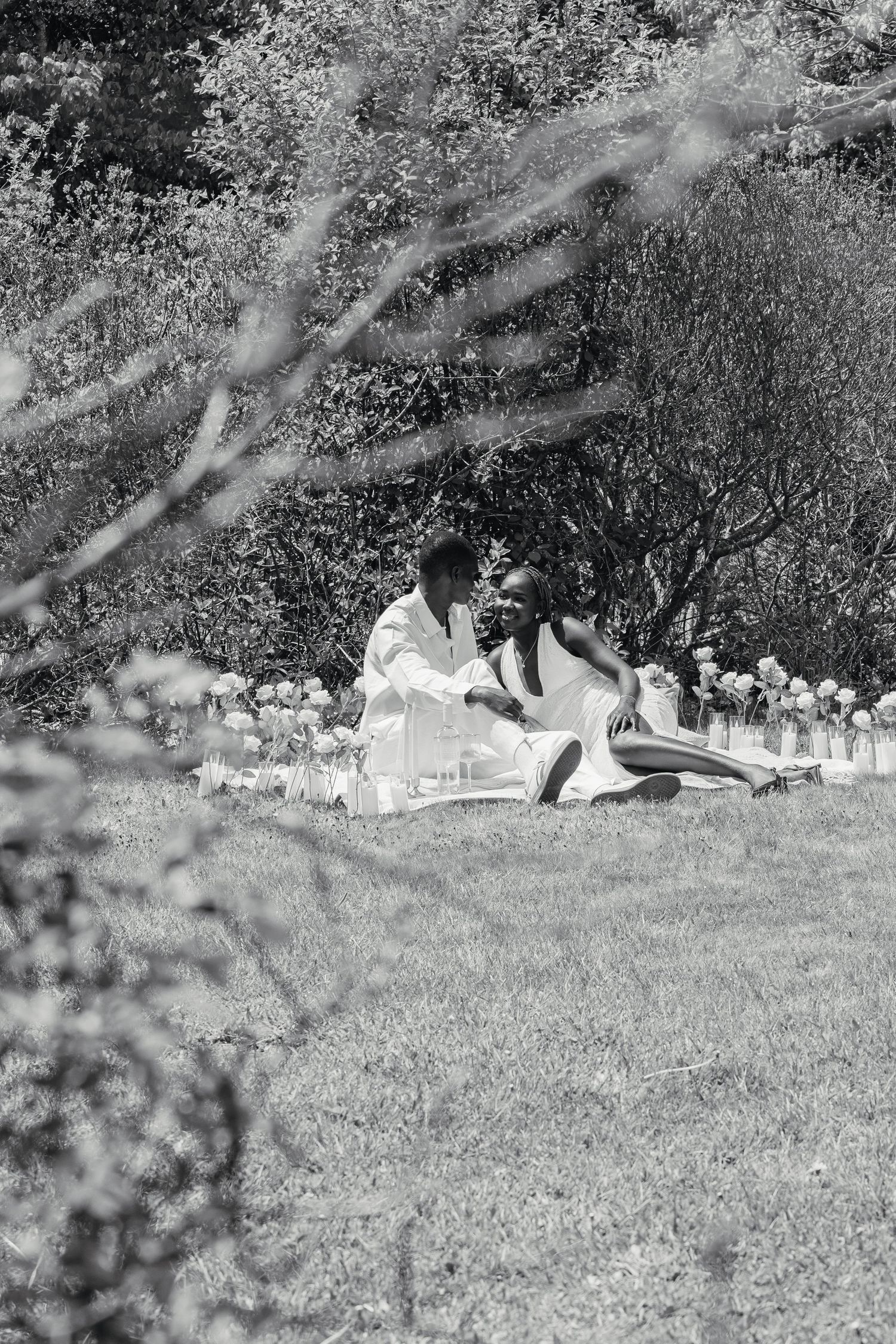Black and white photo of an engaged couple sitting together on grass near floral decorations in a garden.