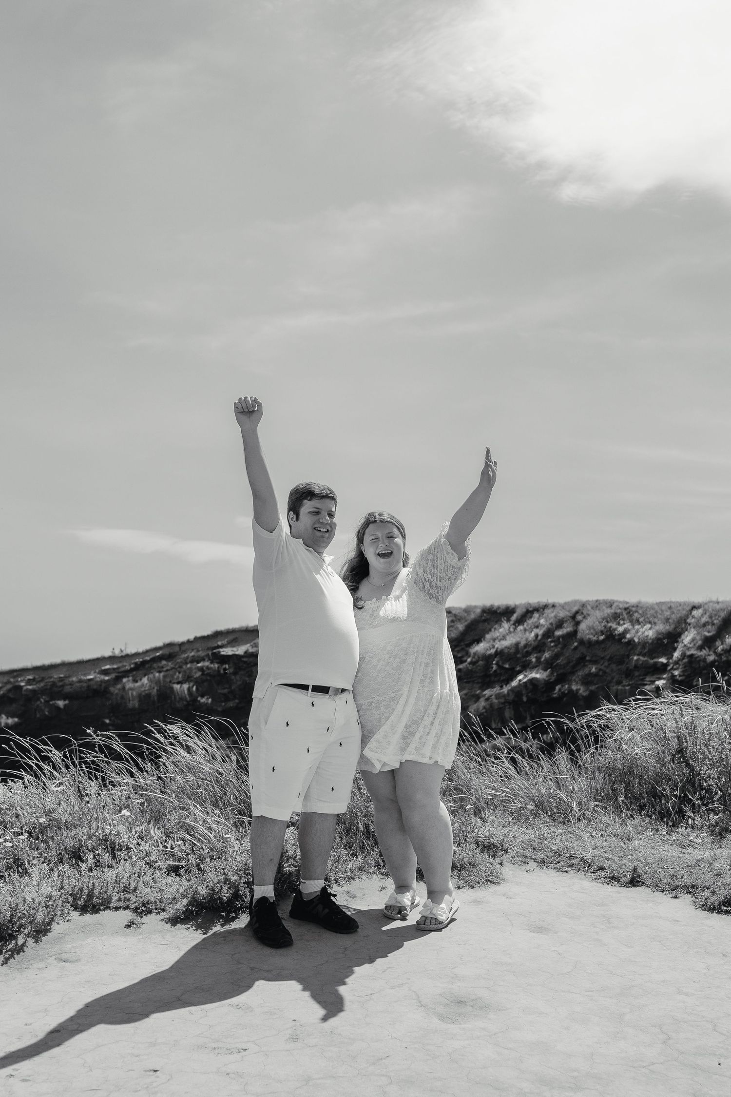Black and white photo of a newly engaged couple celebrating with raised arms on a sunny sandy path.