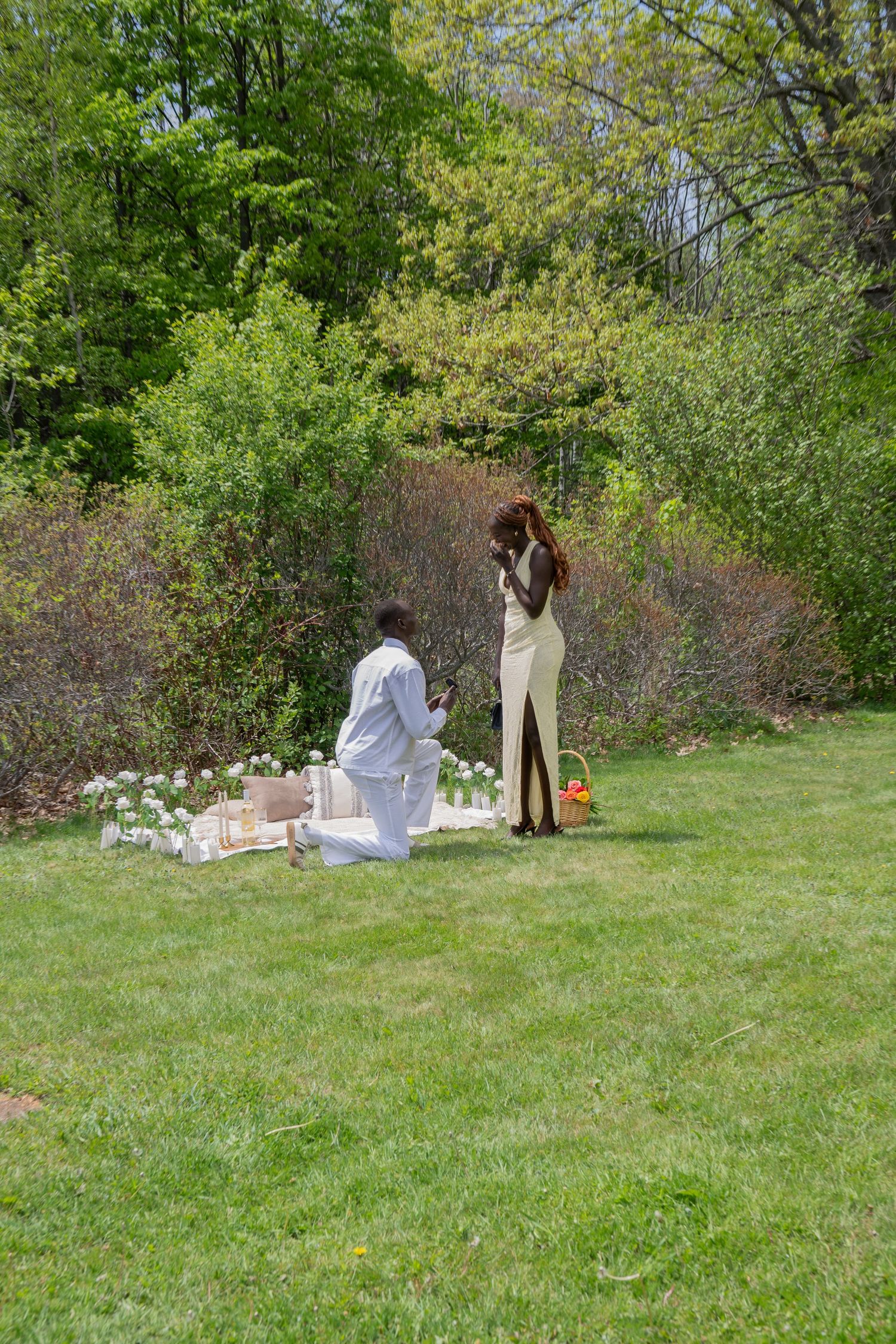 Man kneeling on one knee proposing to woman in gold dress at a decorated outdoor garden picnic.