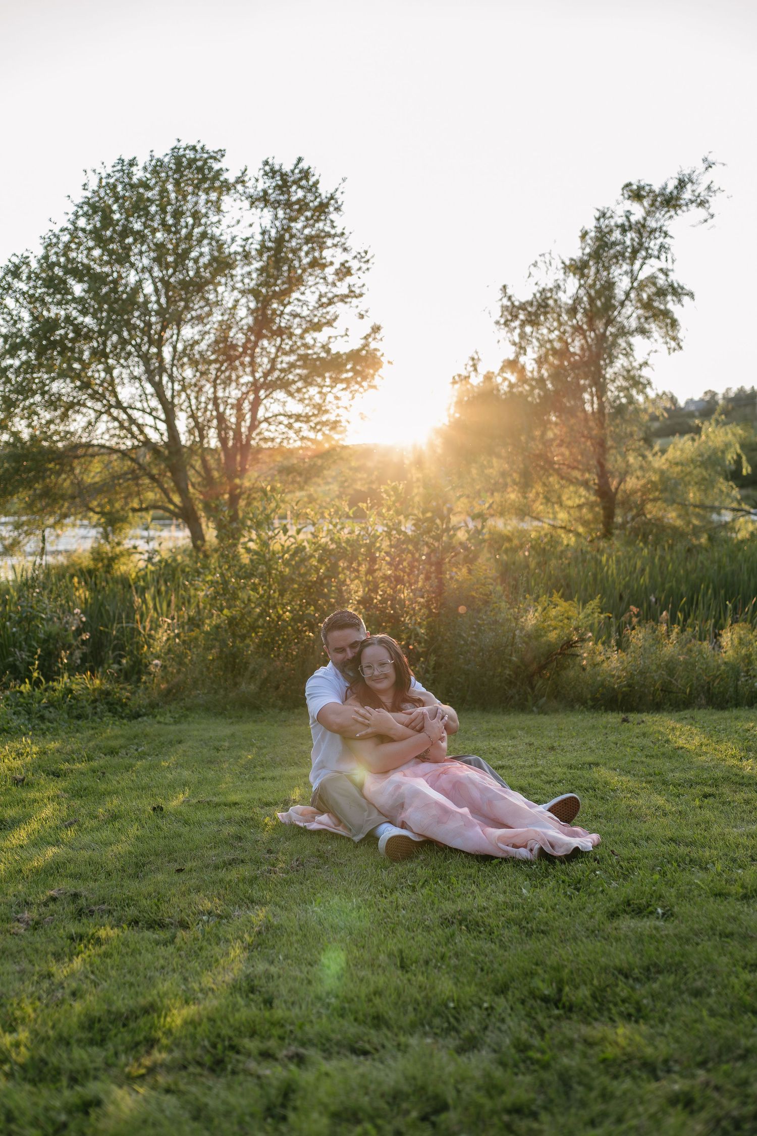 Couple sitting on green grass at sunset, embracing romantically with golden sunlight and trees in background.