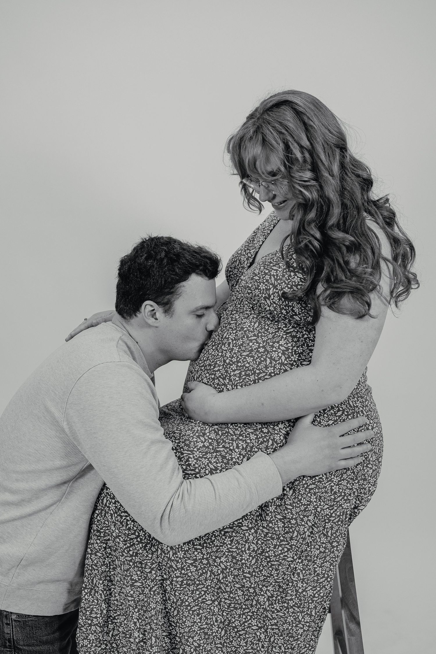 Black and white photo of a man kissing his partners pregnant belly in a studio setting.
