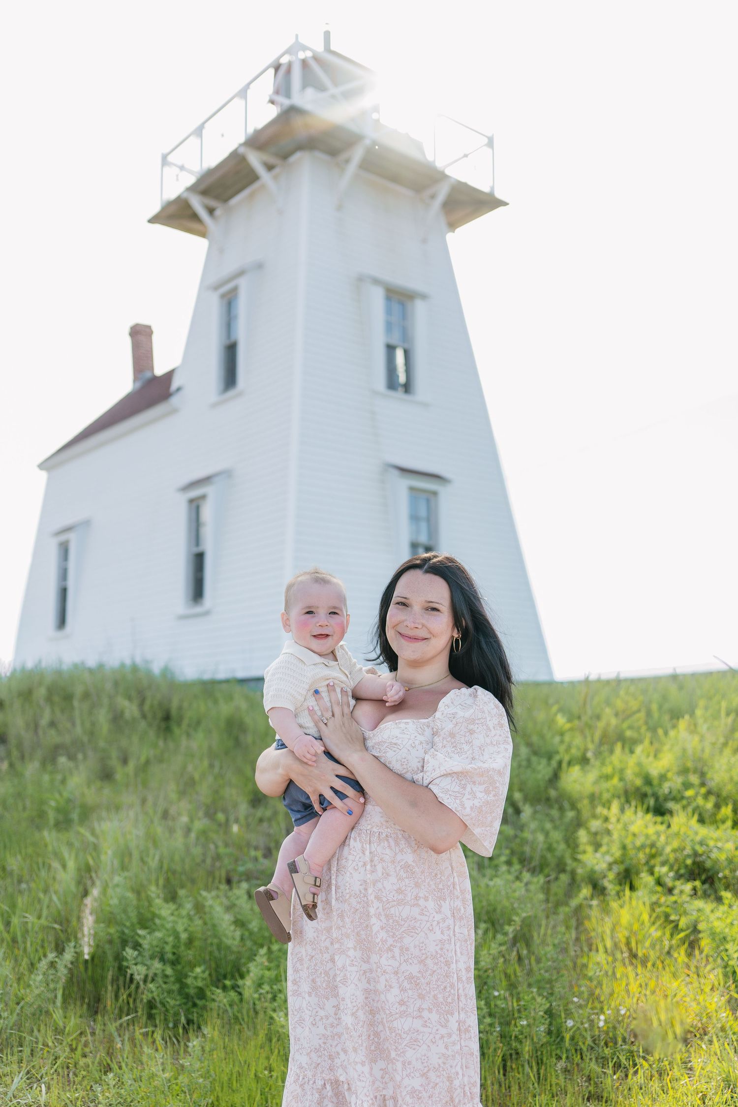 Pregnant woman in floral dress holds baby near white lighthouse on green hillside.