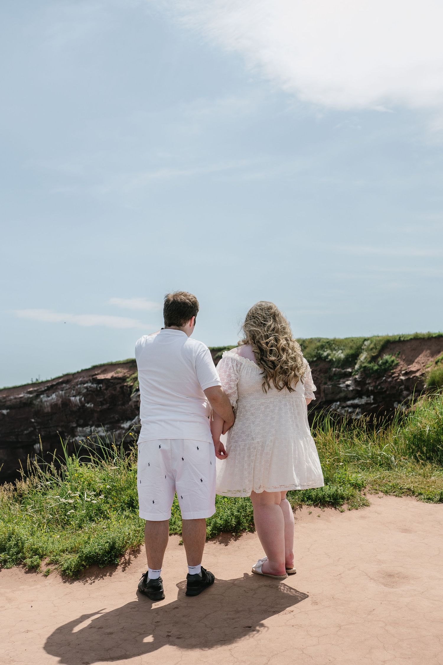 Couple in white outfits holding hands while gazing at scenic coastal cliffs on a sunny day.
