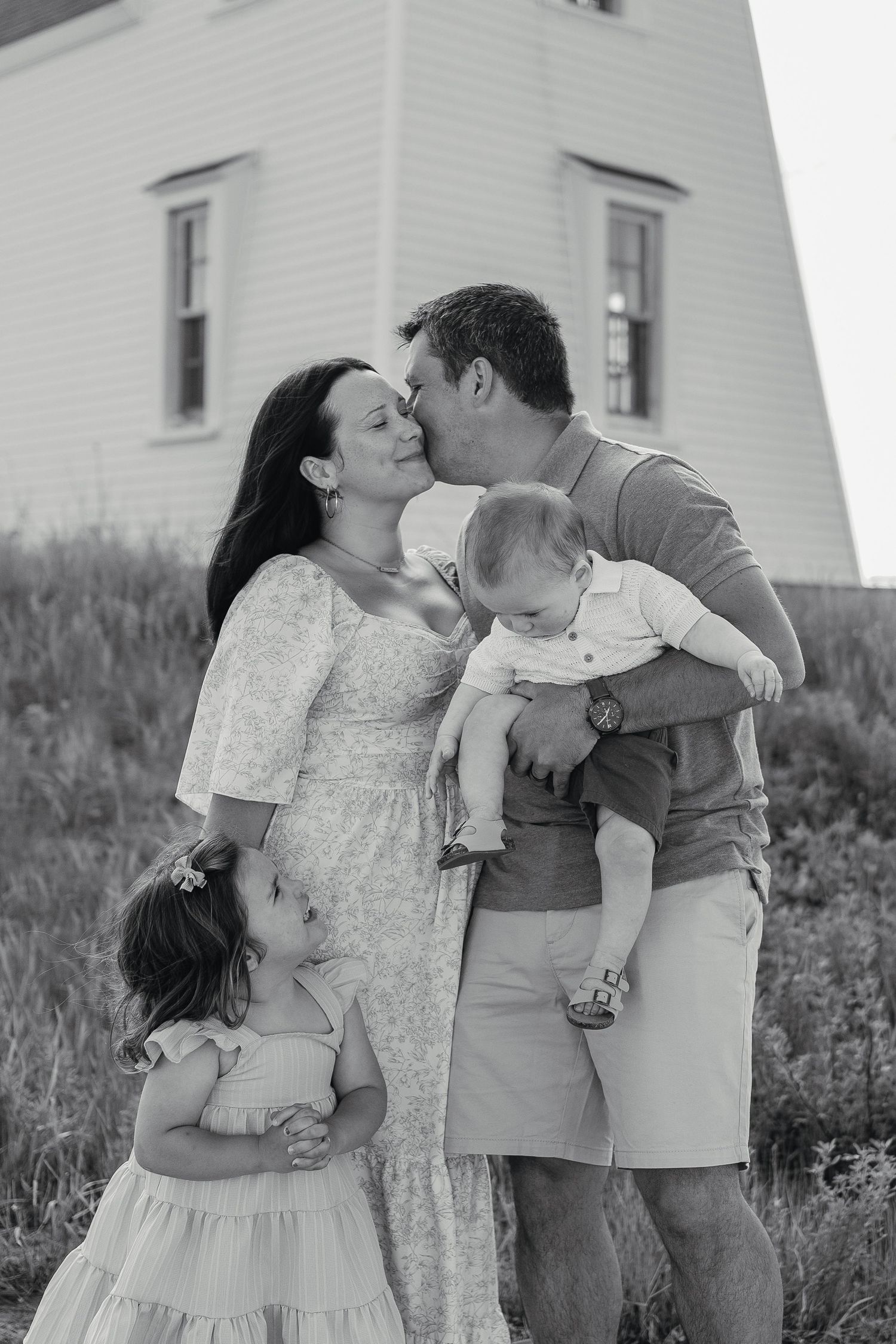 Happy family with two children sharing a kiss outdoors near a white building on a sunny day.