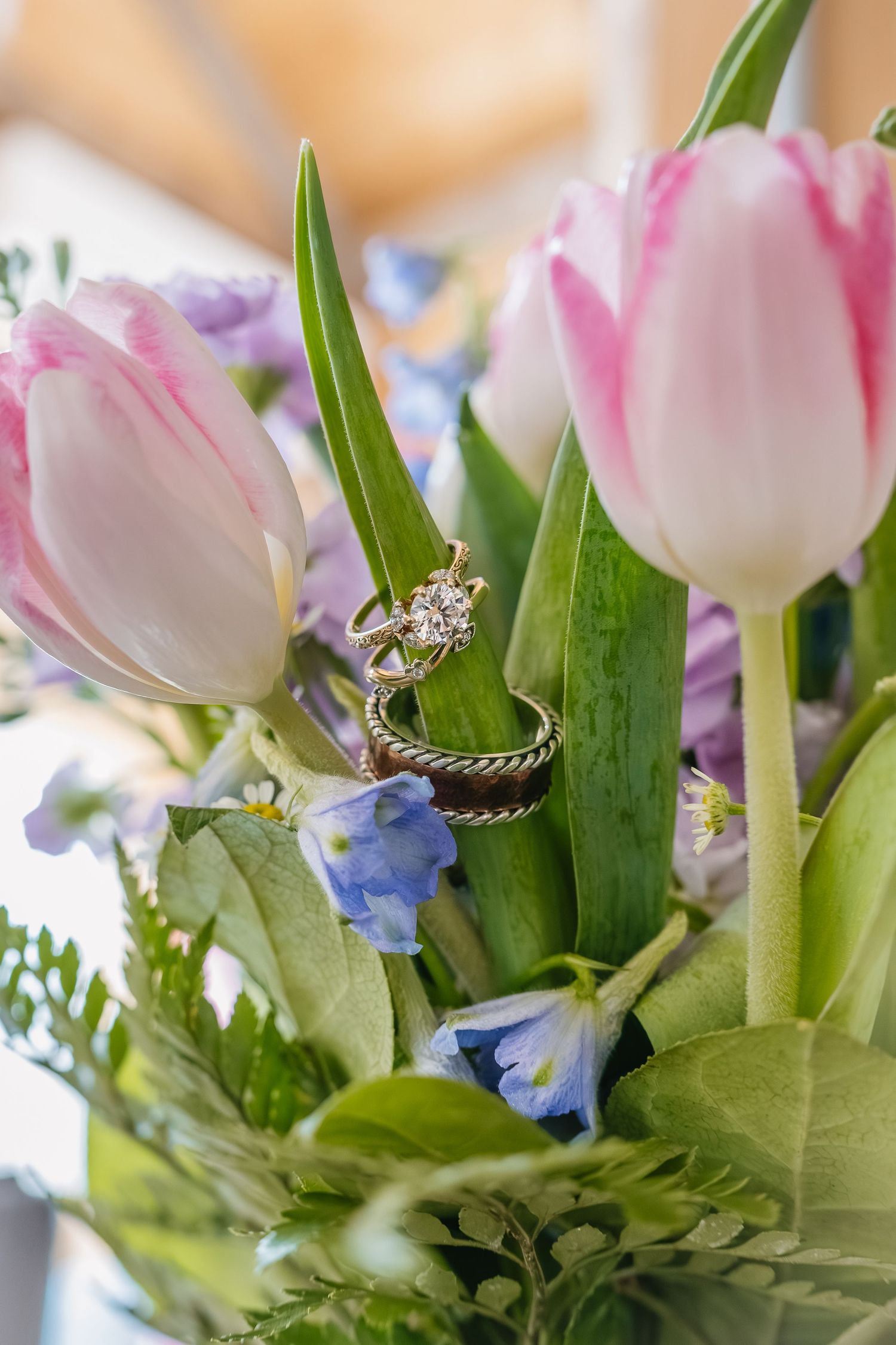 Wedding rings nestled among pink tulips and blue flowers in a bridal bouquet, close-up detail shot.