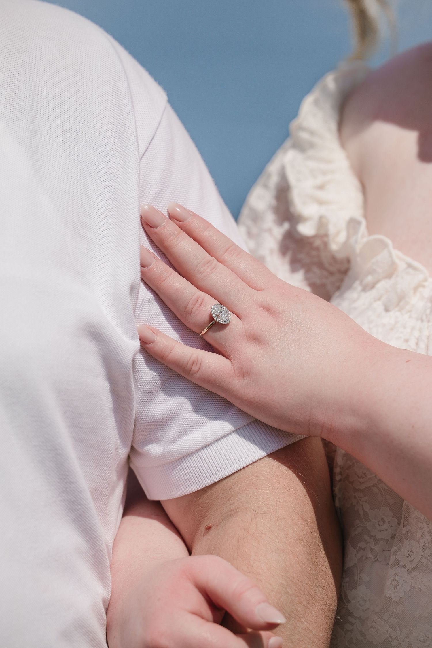 Close-up of a woman's hand wearing a diamond halo engagement ring resting on a man's white shirt.