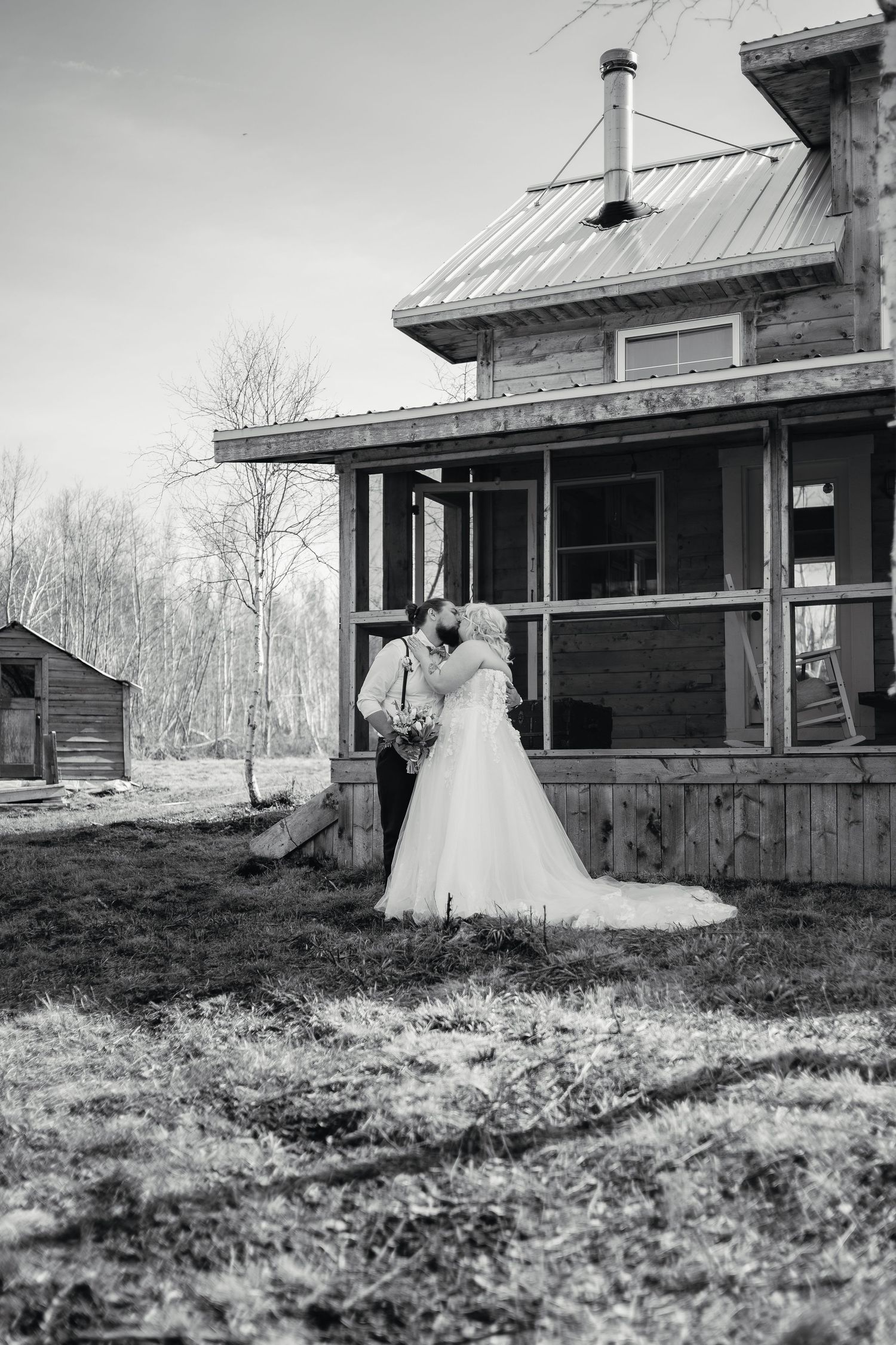 Black and white photo of bride and groom kissing outside rustic wooden farmhouse on wedding day.
